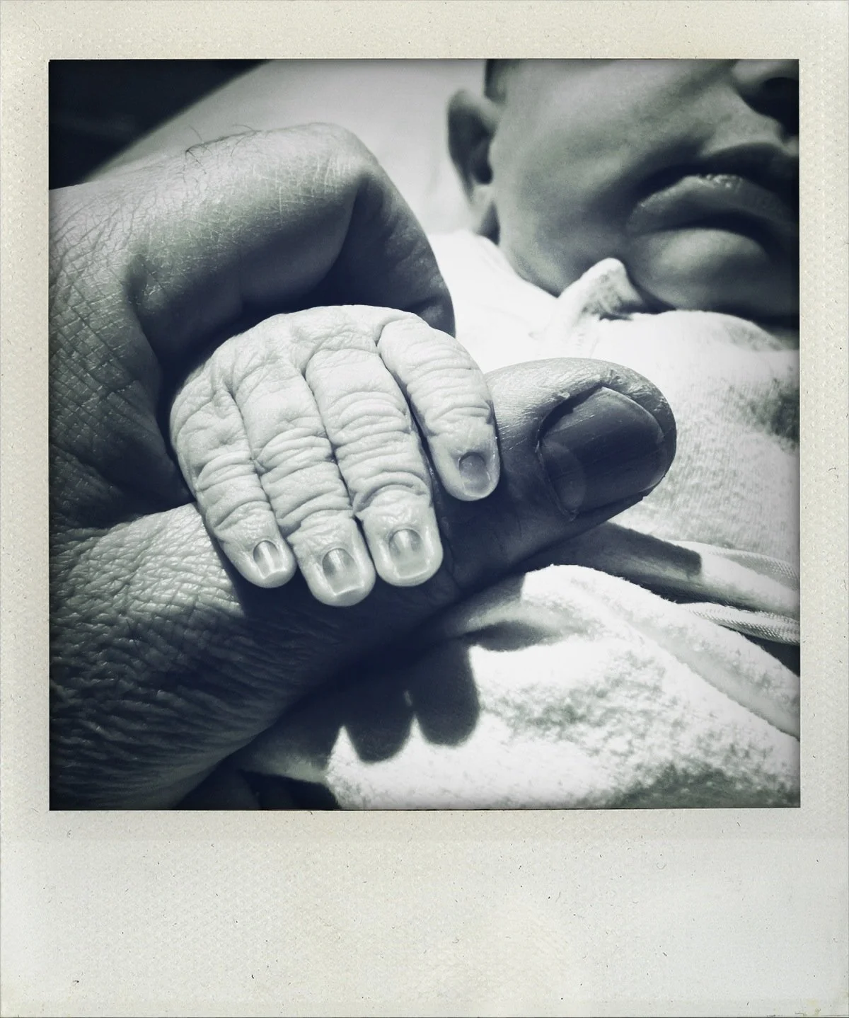Close-up black-and-white photo of a tiny baby's hand gripping an adult's finger, with part of a smiling face visible in the background.
