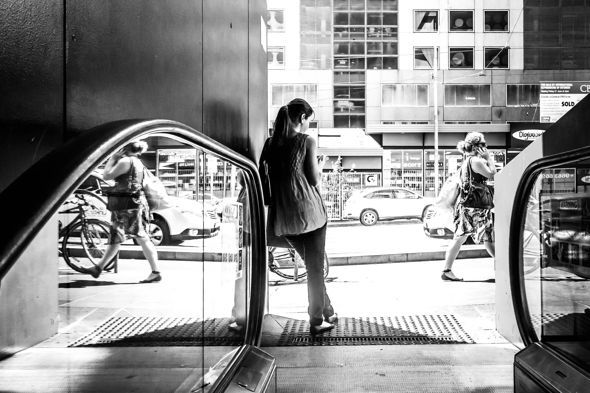 Black and white photo of city street viewed from inside a building, showing two women walking outside, one on each side of the sidewalk, and a young woman standing in the foreground looking at her phone.