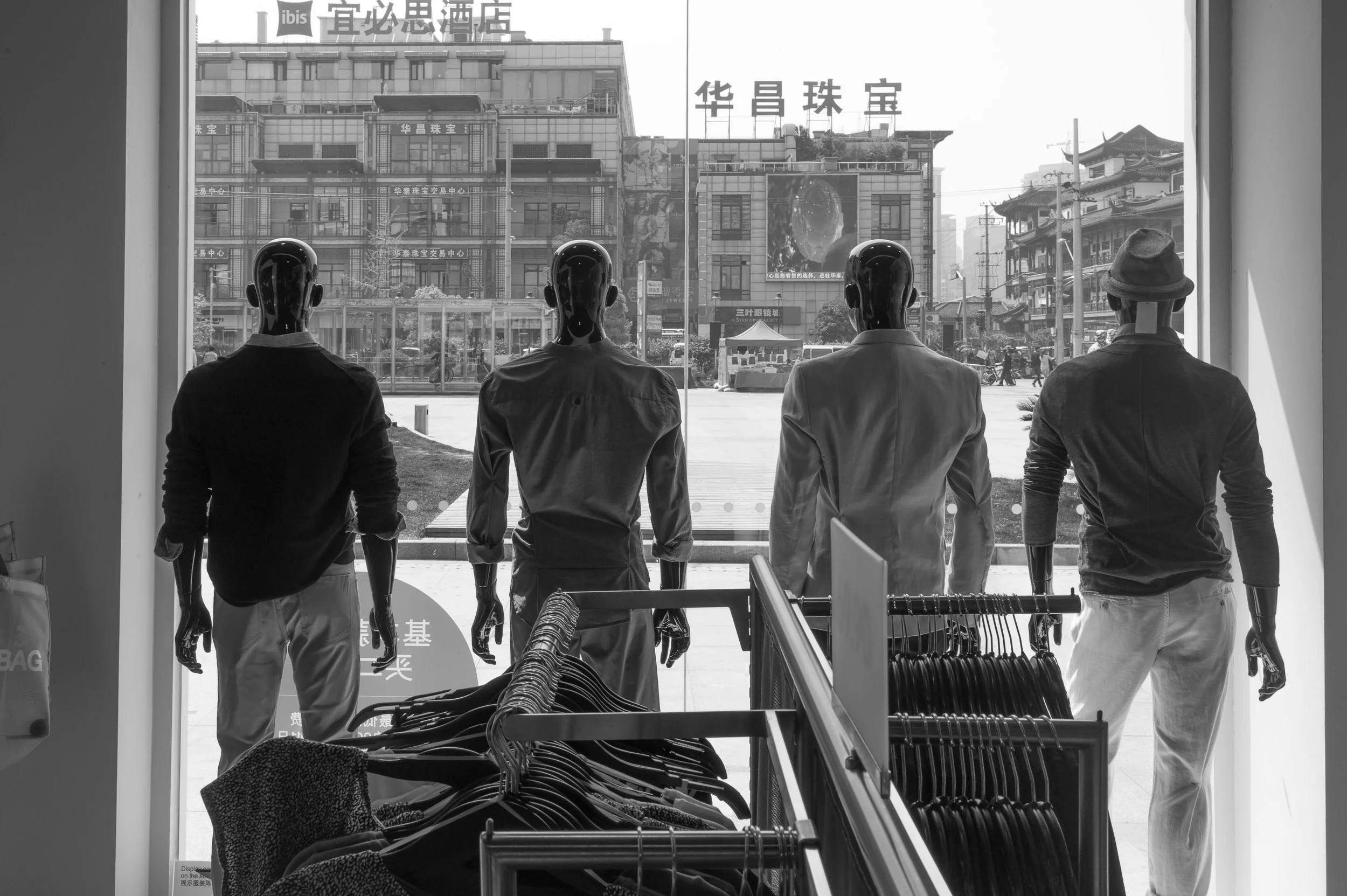 Four mannequins with male features dressed in casual outfits are displayed in a store window, facing outward. Behind them, outside the store, a busy urban street with buildings, signs, and pedestrians is visible.