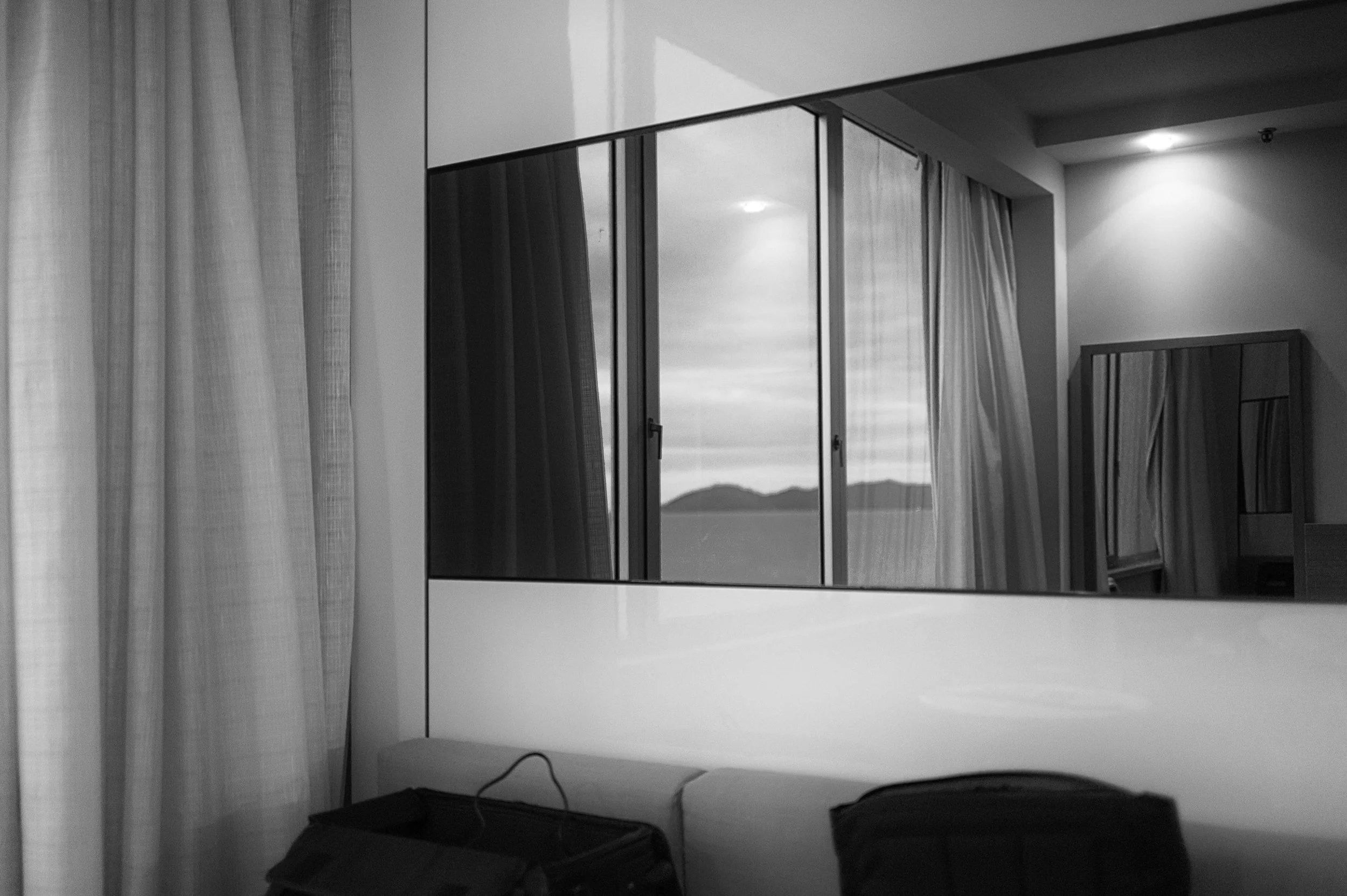 A black and white photo of an interior room showing a window with curtains and mountain scenery outside, a mirror reflecting part of the room, and a bed with a black bag on it.