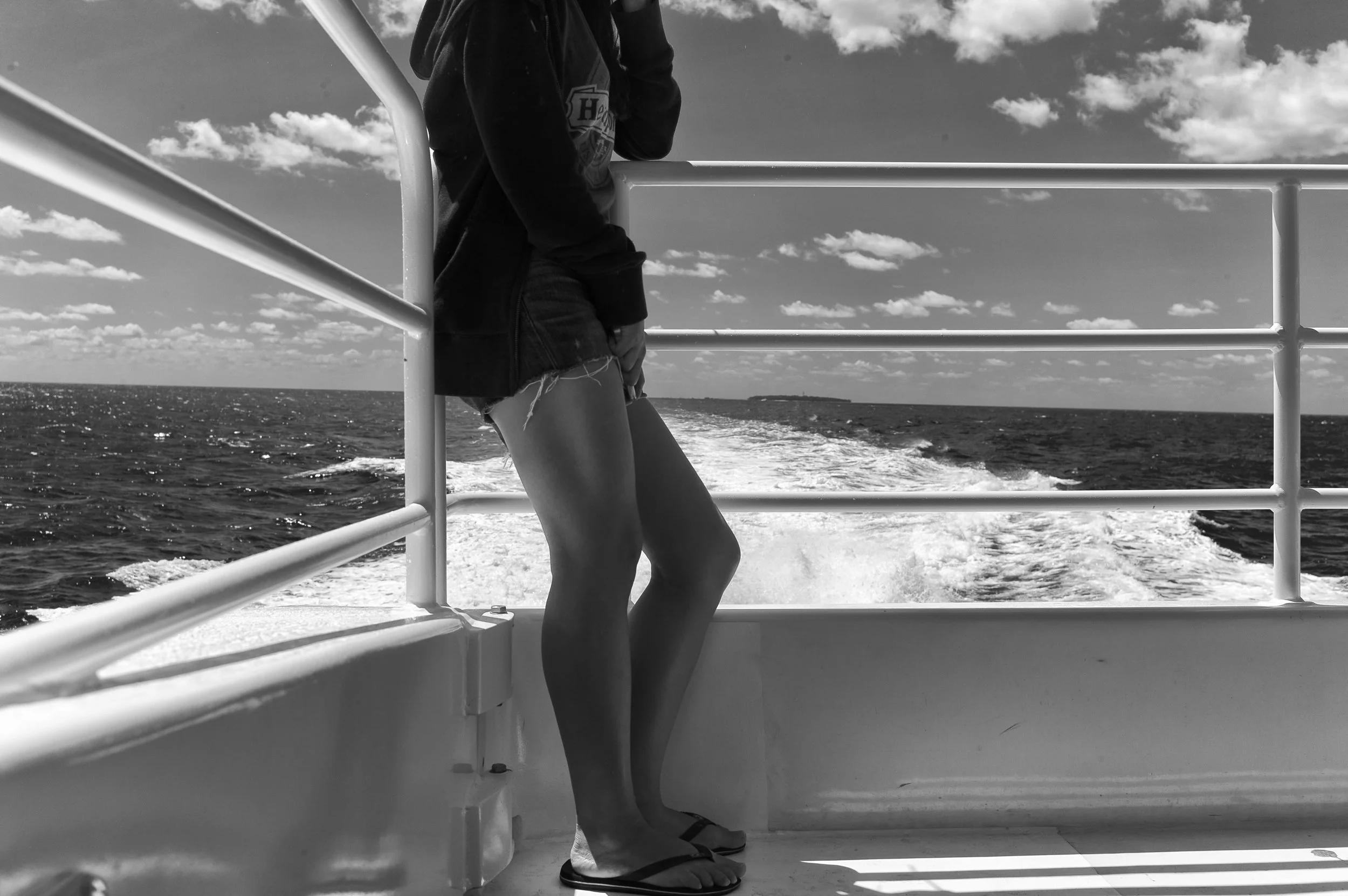 Person standing on a boat, holding onto the railing with the ocean and a partly cloudy sky in the background, in black and white.