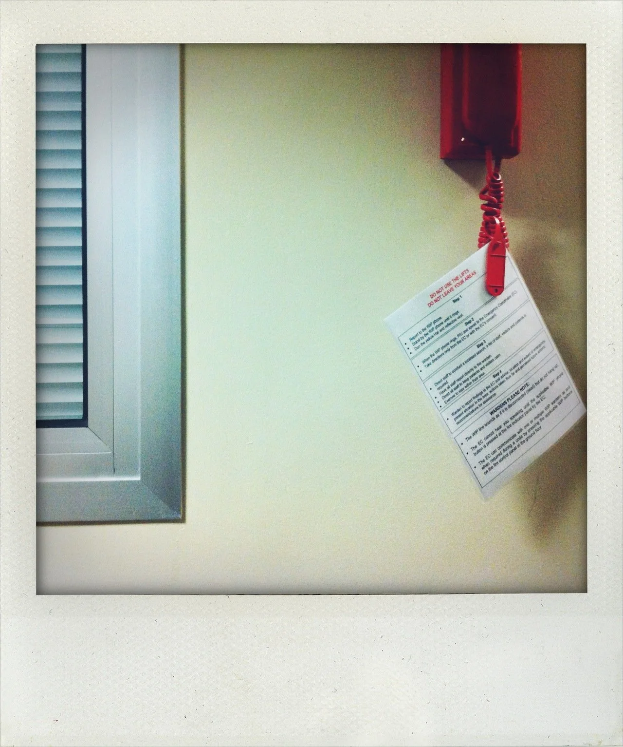 Red fire alarm with a white notification tag hanging from its cord on a beige wall, next to a window with closed blinds.
