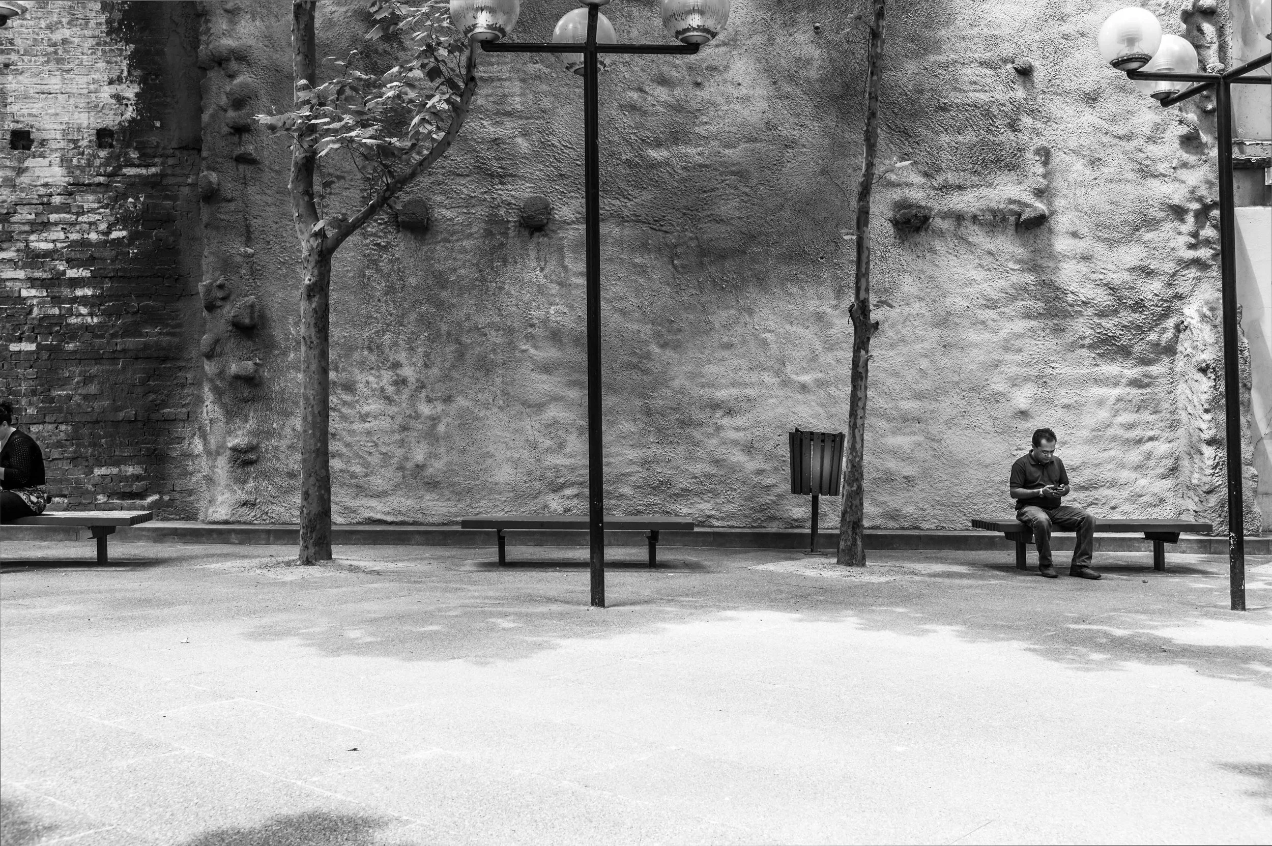 A black and white photo of an urban park with a textured wall, trees, benches, and two people sitting on benches with one person using a phone.