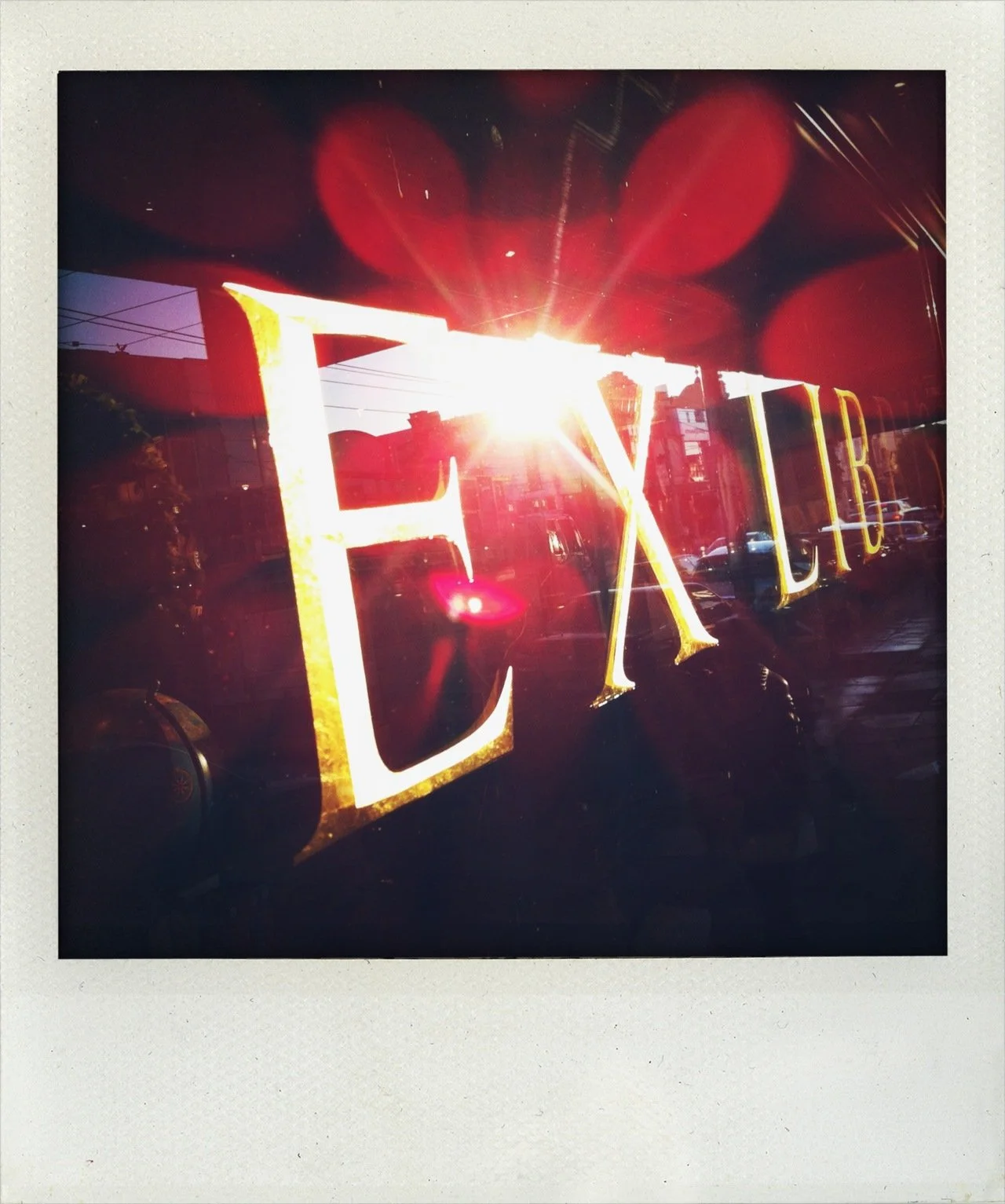 Close-up of a window with a neon sign that reads 'EXPL' illuminated, sunlight shining through the sign creating glare and reflections, with a reflection of nearby cars and power lines in the glass.