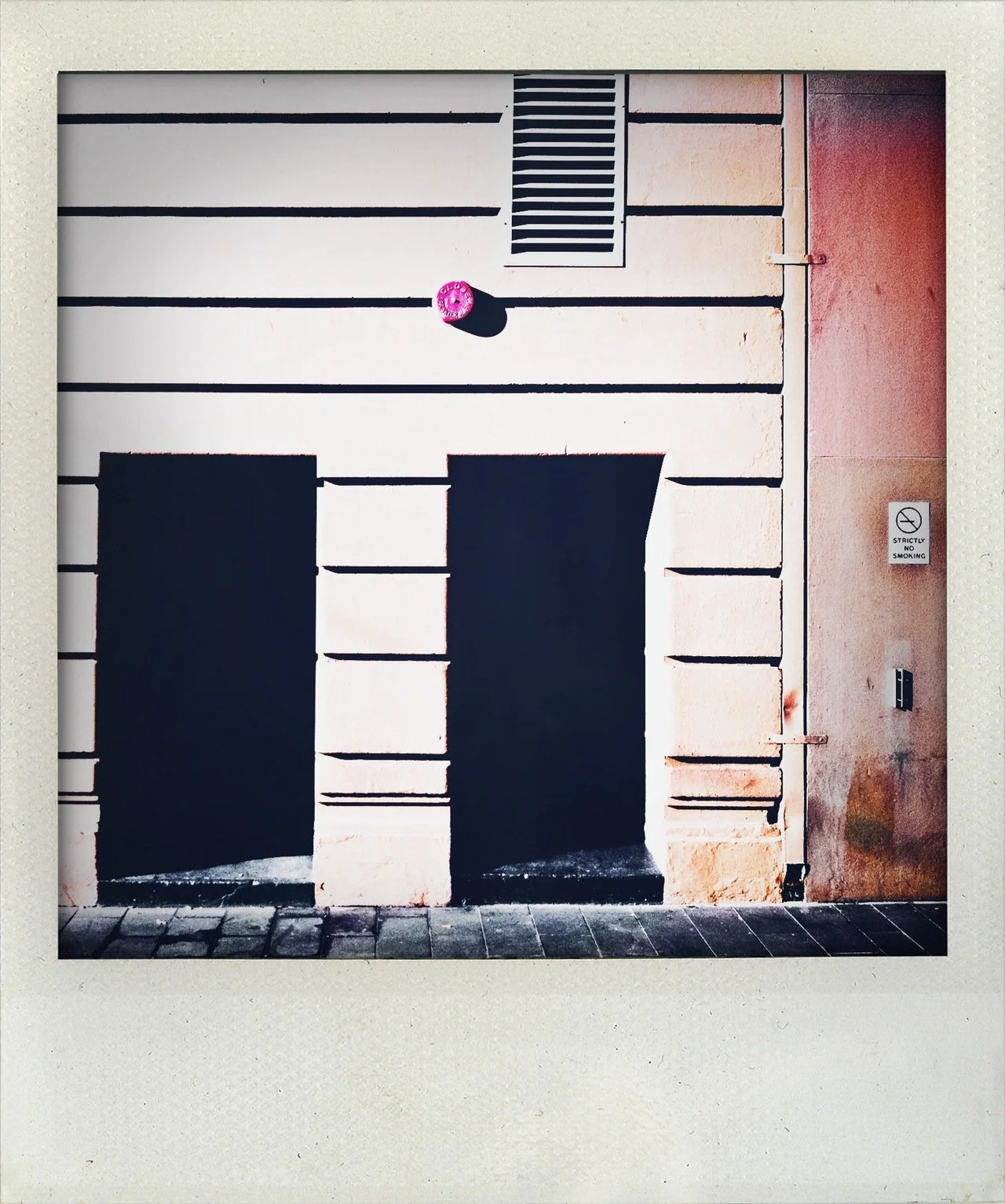 Polaroid photo of a building door with black rectangles painted to resemble a face, with a small pink object above the door and a "No Smoking" sign on the right side.