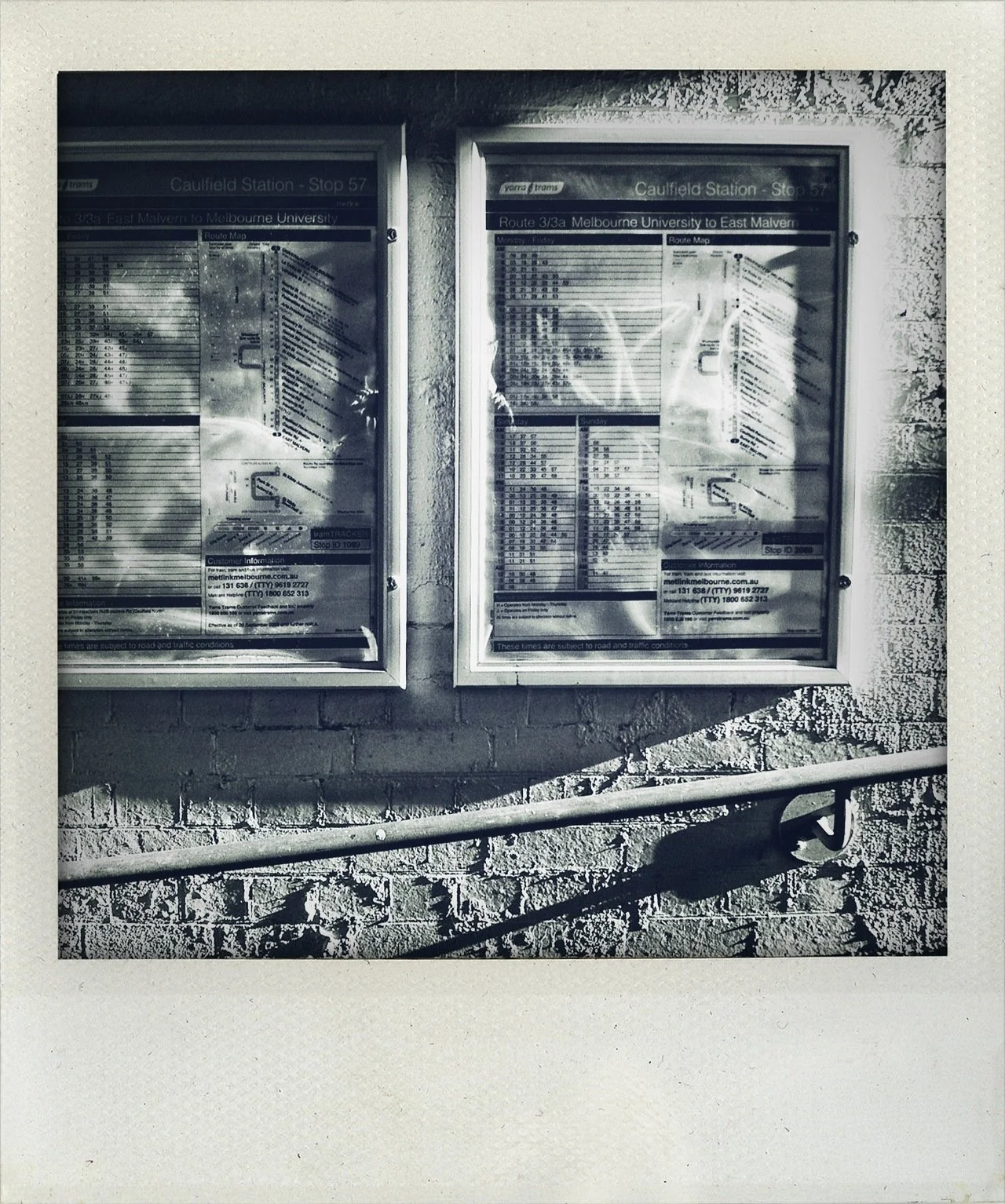 Two bus schedule display boards at Caulfield Station, showing route information for Route 3/3a from Melbourne University to East Malvern, mounted on a brick wall with a metal handrail below.