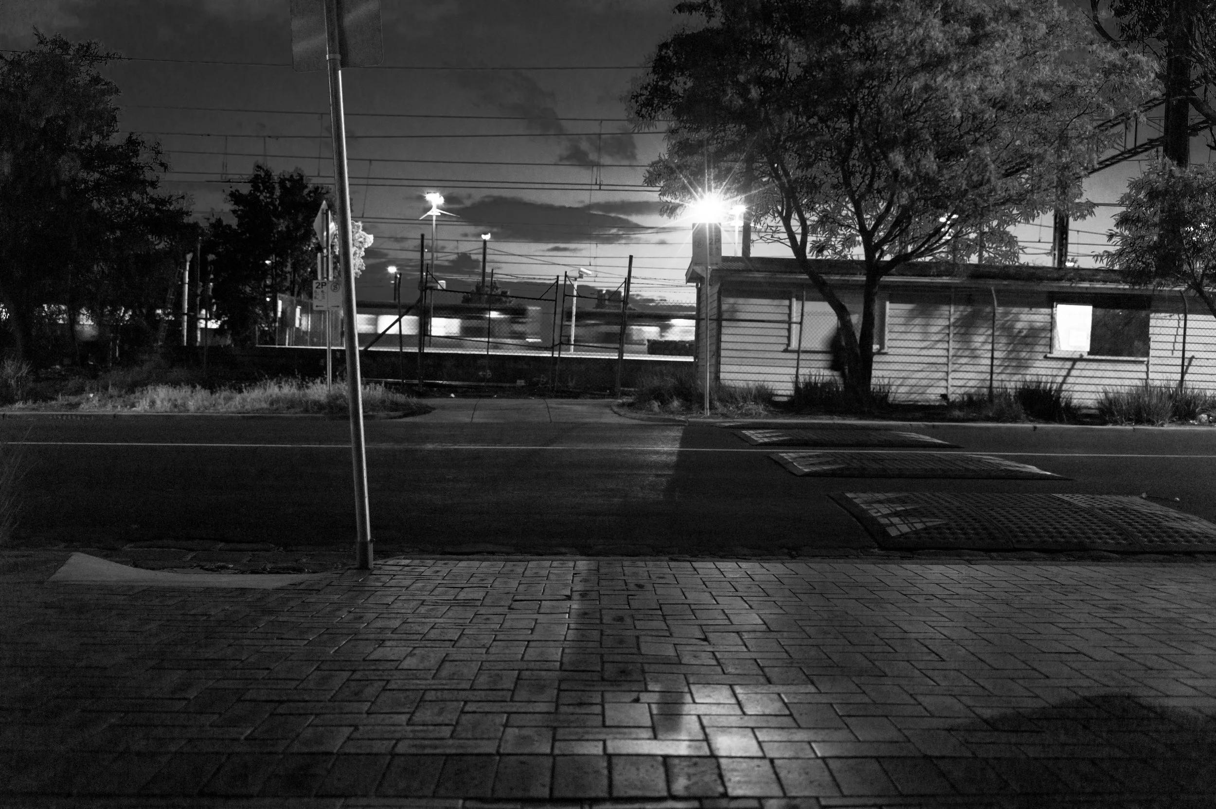 A black and white photo of a night scene with a pavement foreground, a street, and a residential building behind a fence. There are trees, power lines, and bright streetlights illuminating the area. The sky is partly cloudy.