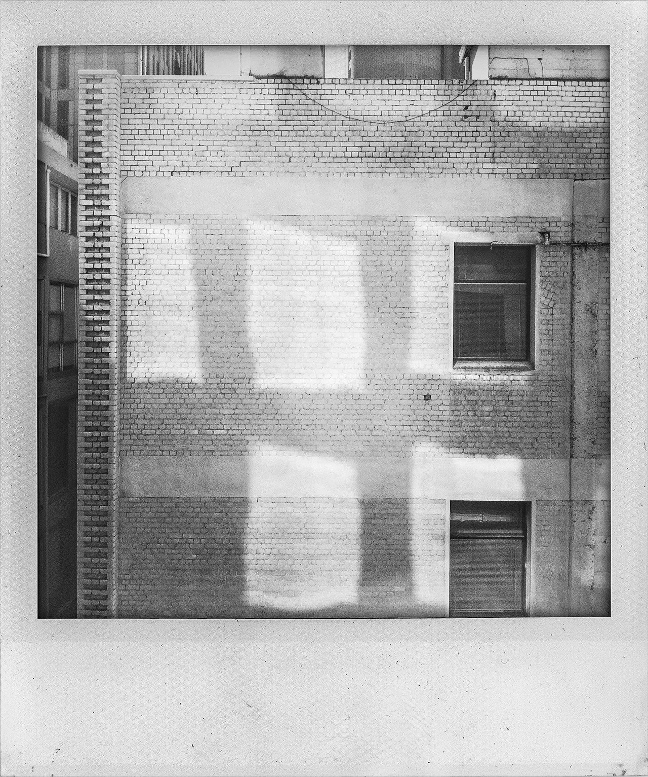 Black and white photo of a brick building's exterior wall with two windows and shadow patterns of window frames cast on the wall.