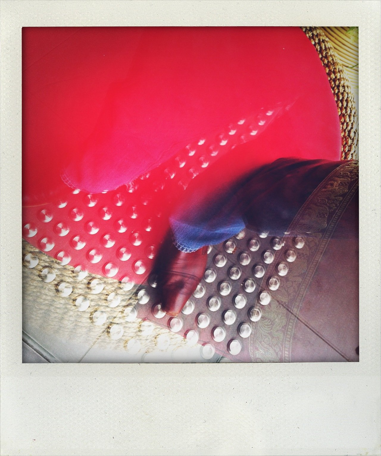 A person's foot wearing a shoe, stepping onto a vibrating massage plaque with white rounded bumps, next to a pink massage cushion with similar bumps, on a tiled floor.