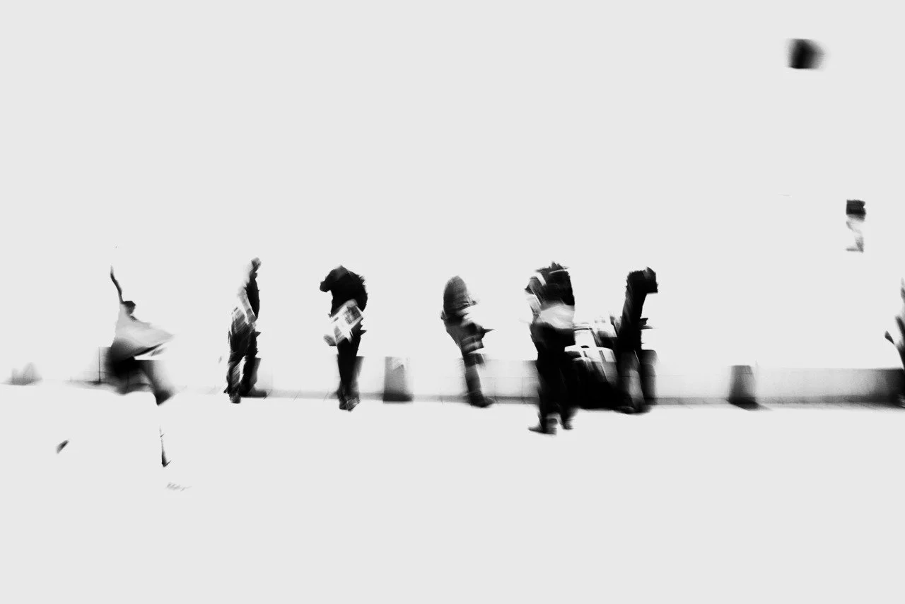 Blurred black and white photo of people standing in a line along a sidewalk, some with luggage or bags.