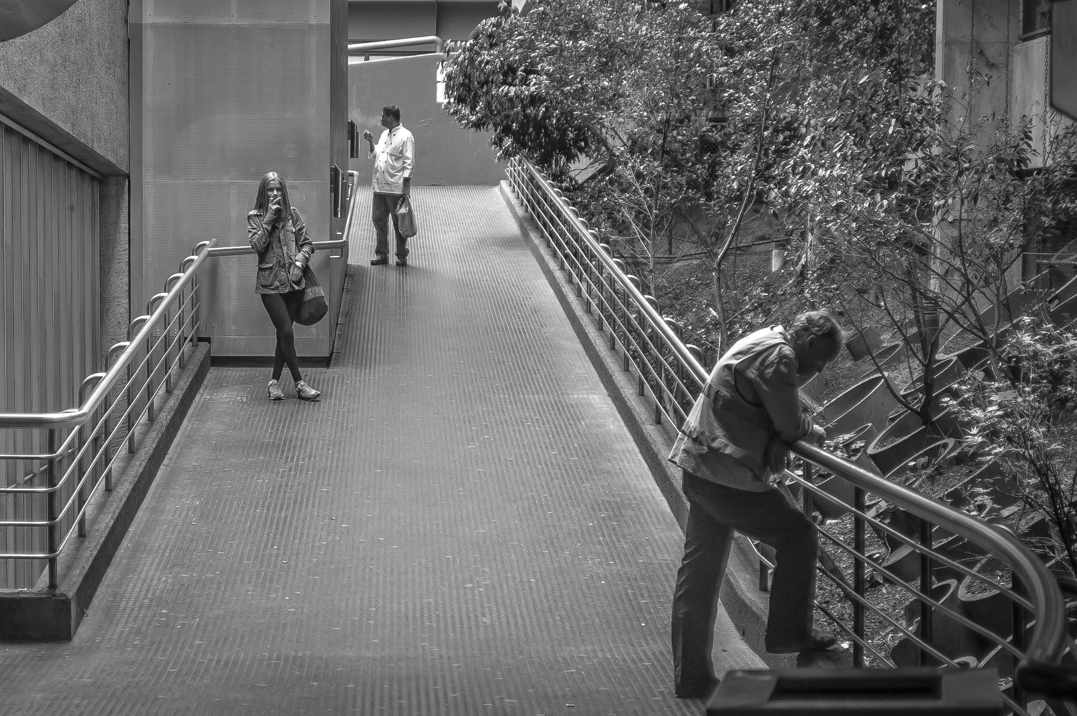 Black and white photo of people on a walkway, with some talking on their phones and one person leaning on the railing, surrounded by trees and building structures.