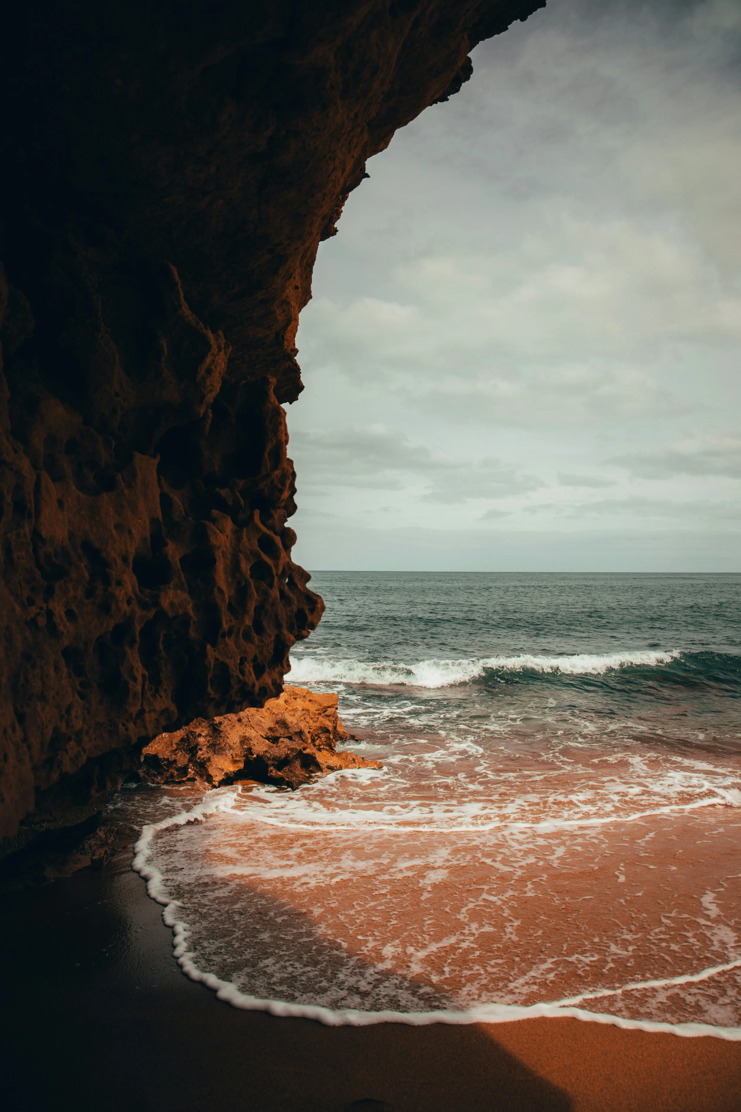 View of a rocky cliffside overlooking the ocean with waves crashing on the sandy beach under a cloudy sky.