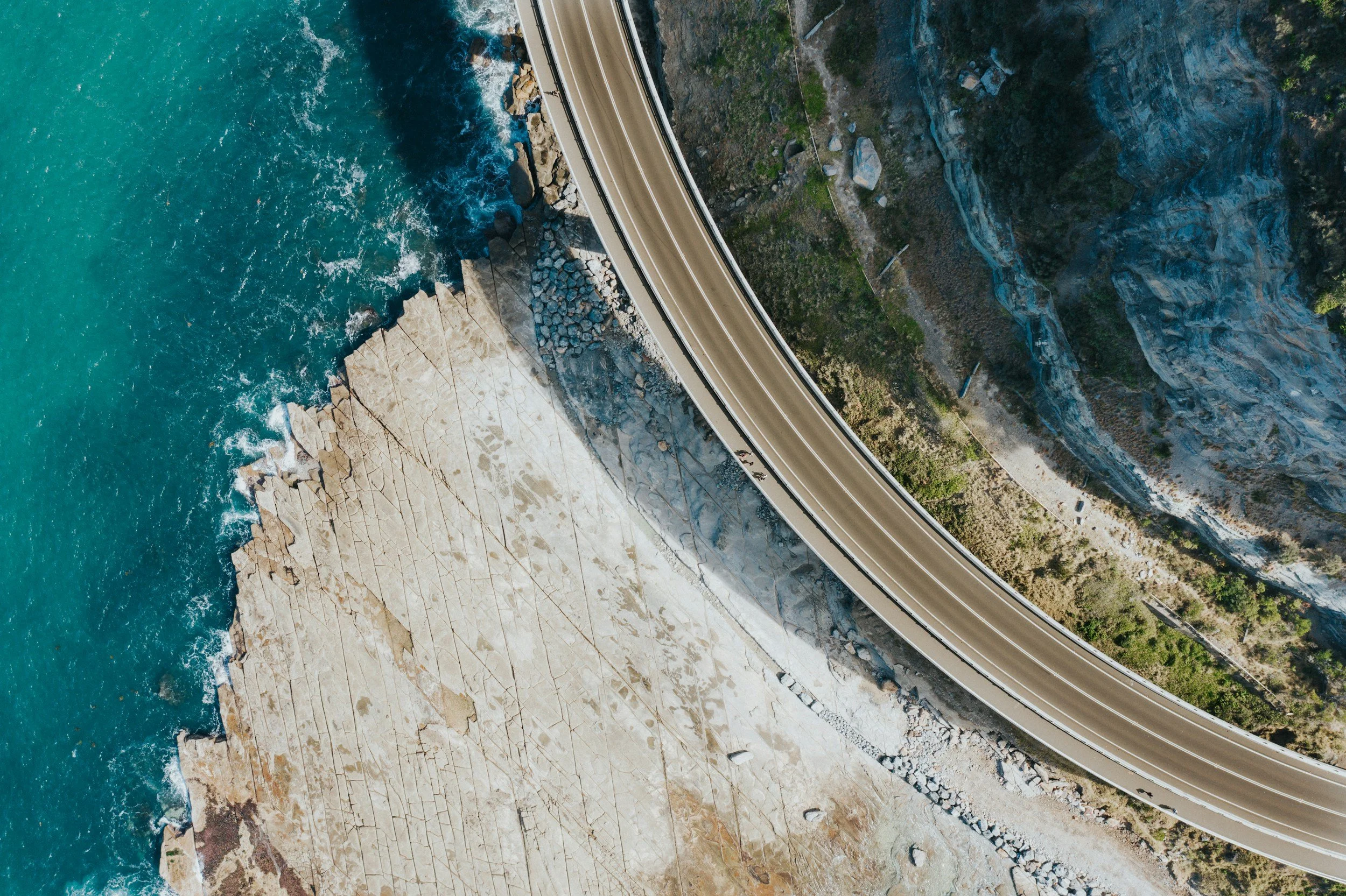 Aerial view of a coastal road running along rocky cliffs next to a turquoise ocean with waves crashing against the rocks.