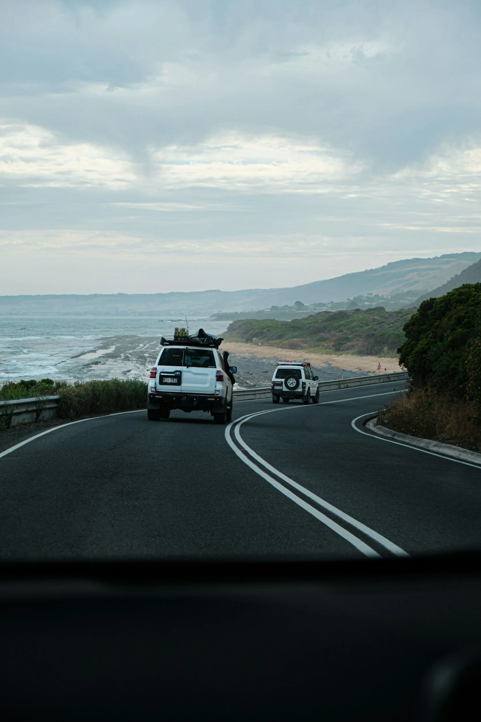 A view from inside a vehicle looking at a coastal road with two parked off-road vehicles, near the ocean with waves and hills in the background, and an overcast sky.