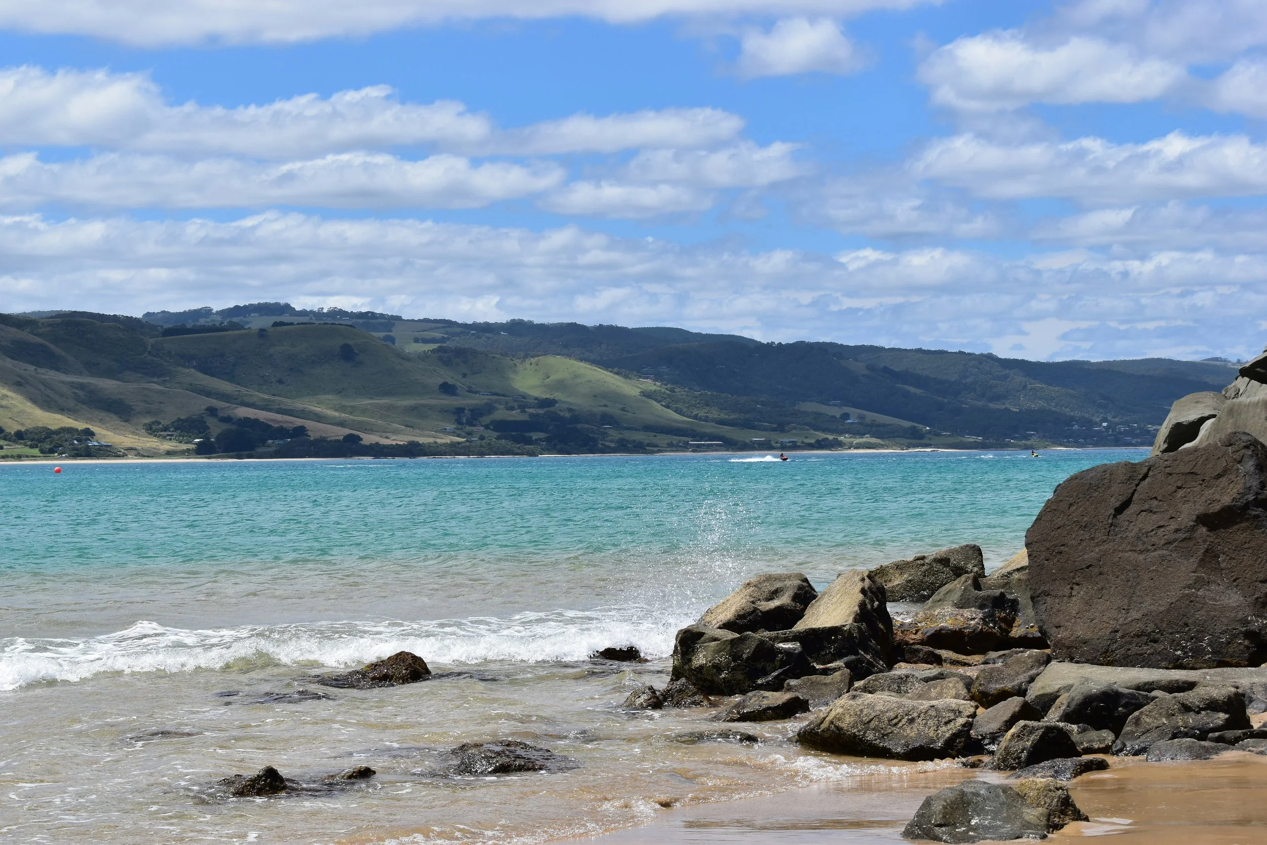Scenic view of a beach with rocks in the foreground, calm turquoise water, green hills in the background, and a partly cloudy sky.