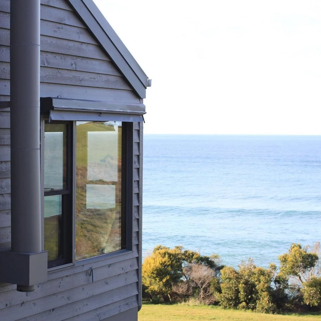 A house with wooden siding and a large window overlooking the ocean, with trees and grassy area in the foreground.