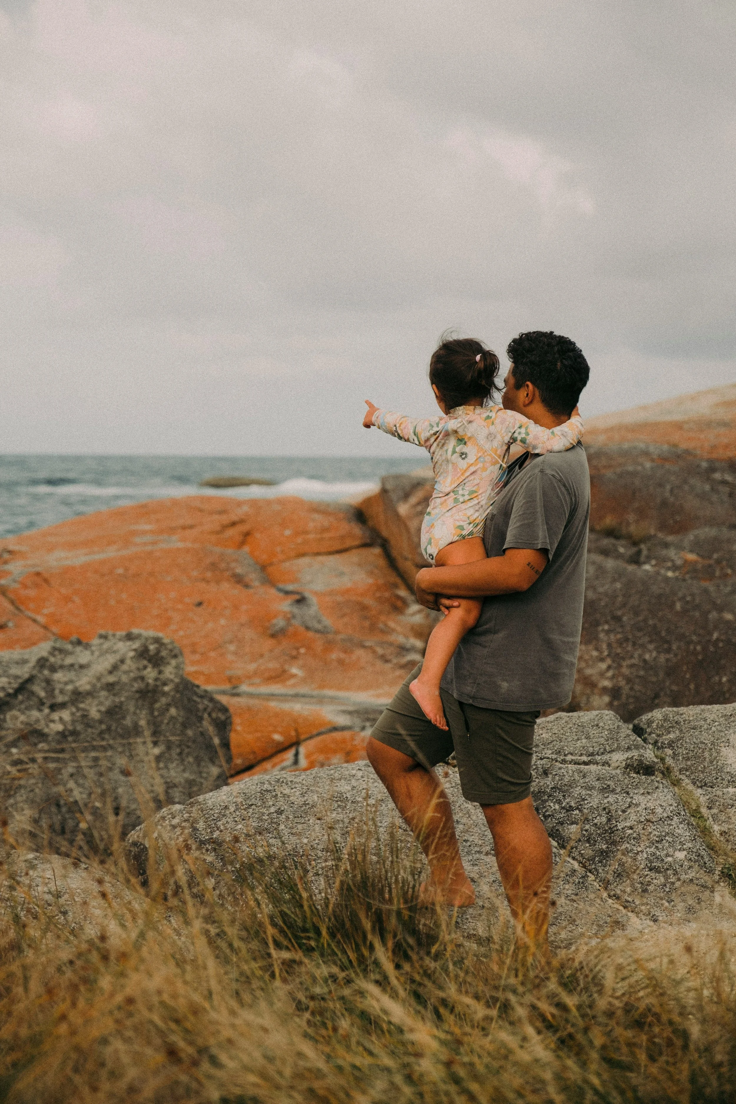 A man holding a young girl in his arms on a rocky beach, with the girl pointing towards the ocean and sky, overcast weather.