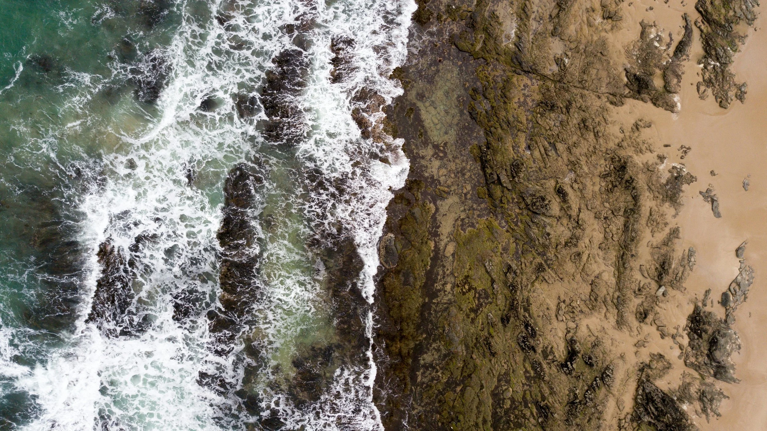 Aerial view of a rocky shoreline with waves crashing onto the rocks and sandy beach.