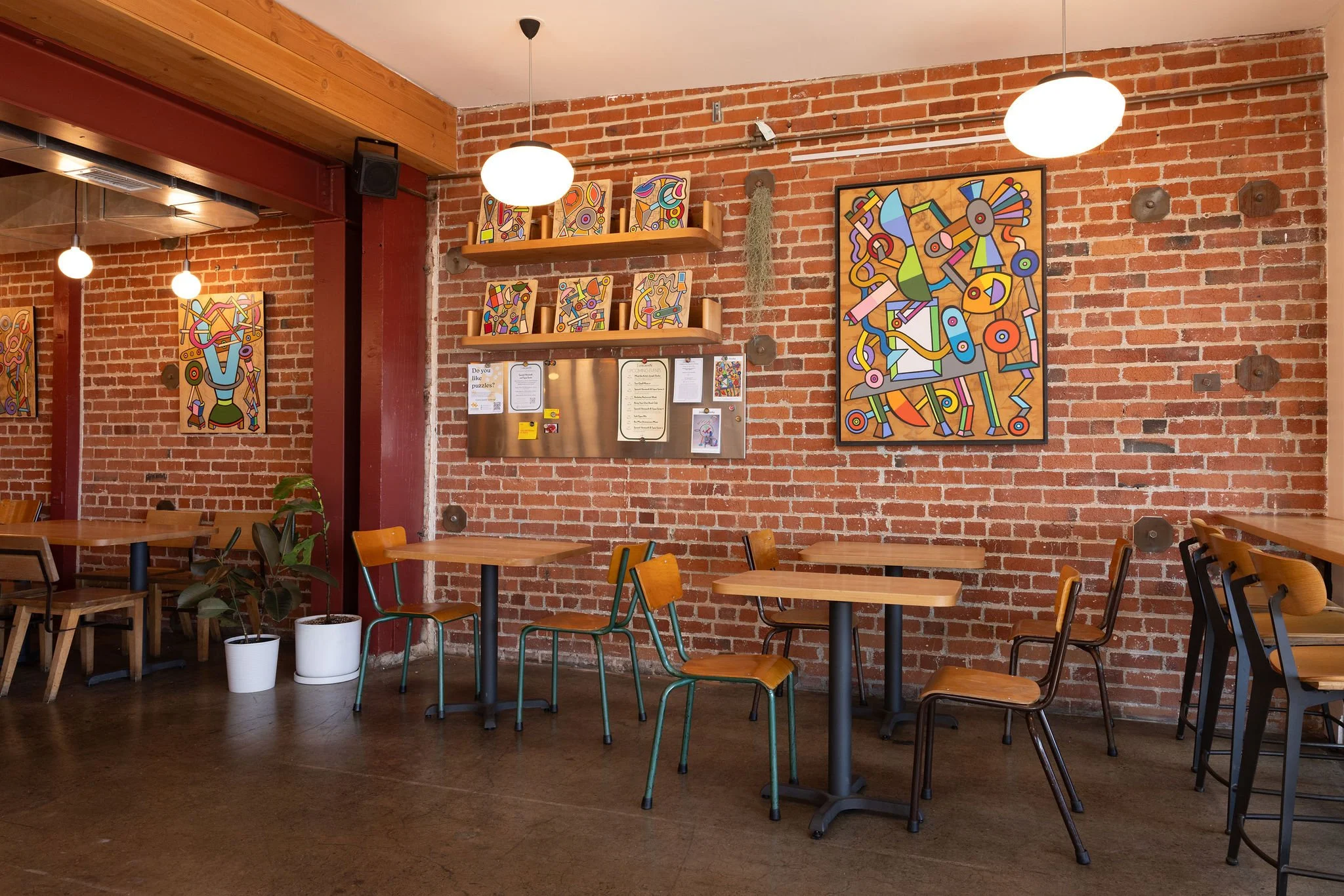 Evergreen Cafe_Berkeley, CA_Inside a cafe with brick walls, wooden tables, and chairs, decorated with colorful abstract art on the walls and hanging ceiling lights.
