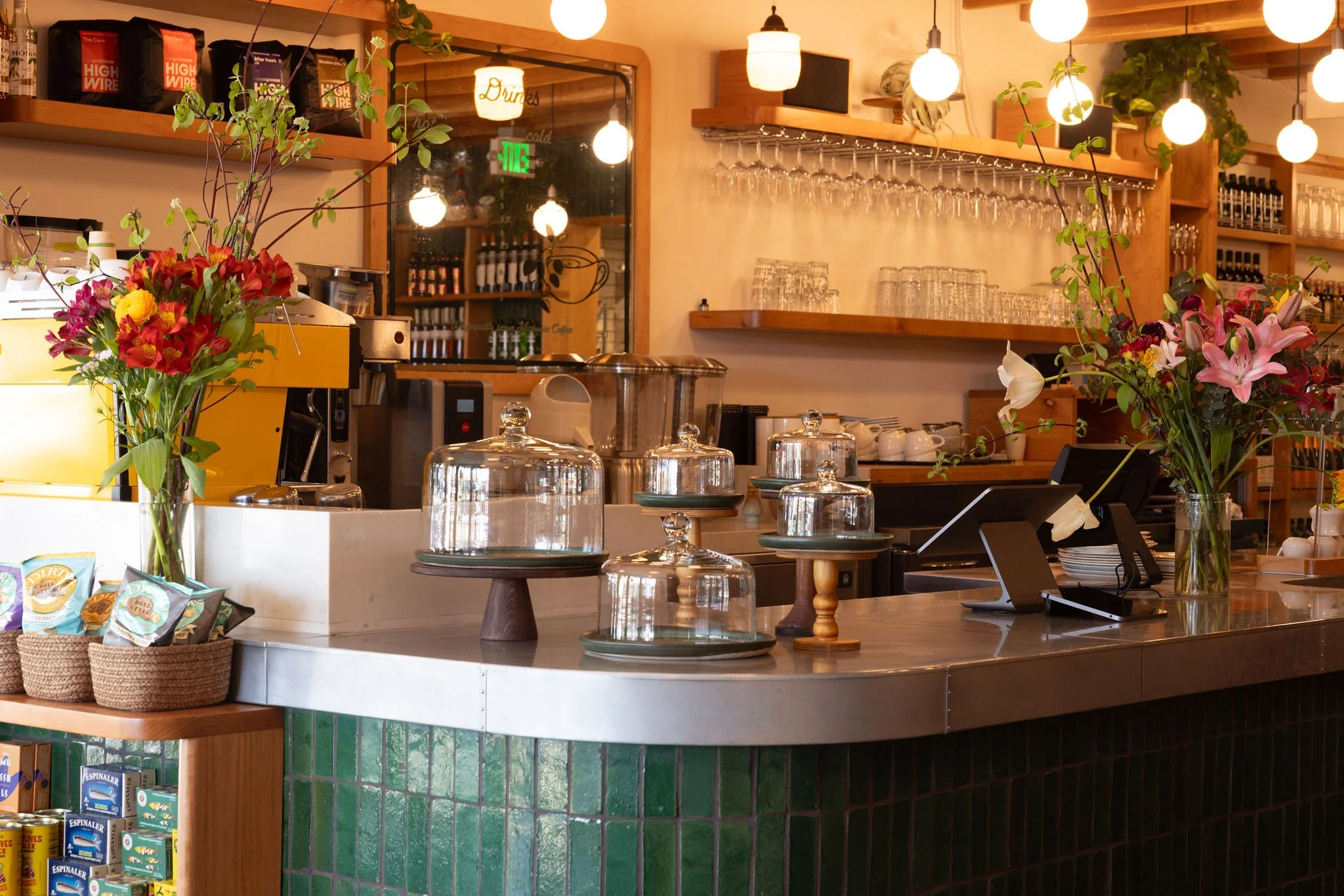 Evergreen Cafe_Berkeley, CA_Interior of a cozy cafe featuring a counter with flower arrangements, glass cake stands, and a coffee machine, with warm lighting and wooden shelves stocked with glasses and bottles in the background.