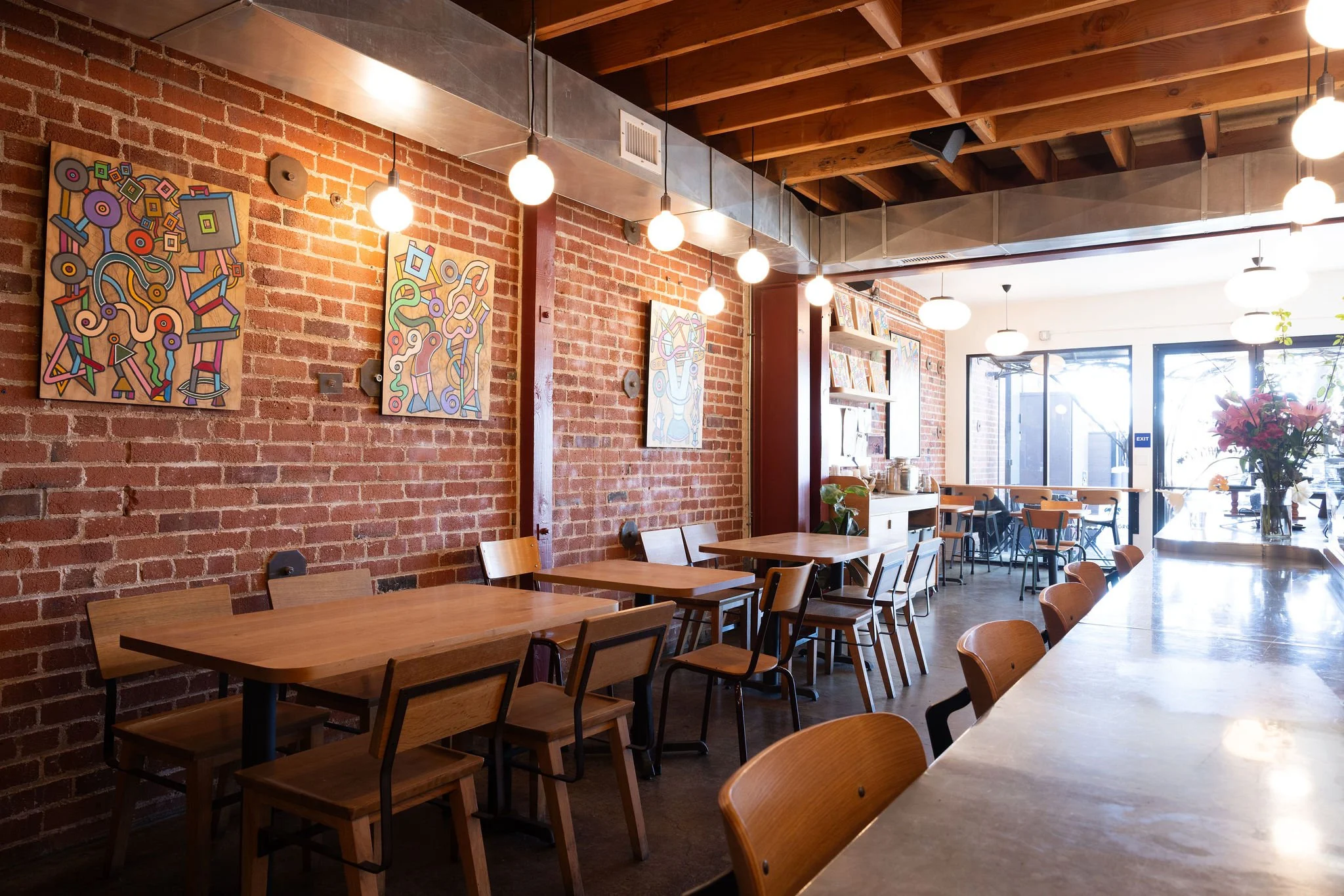 Evergreen Cafe_Berkeley, CA_Interior of a cozy cafe with wooden furniture, exposed brick walls decorated with colorful abstract artworks, hanging ceiling lights, large windows letting in natural light, and a vase of flowers on the counter.