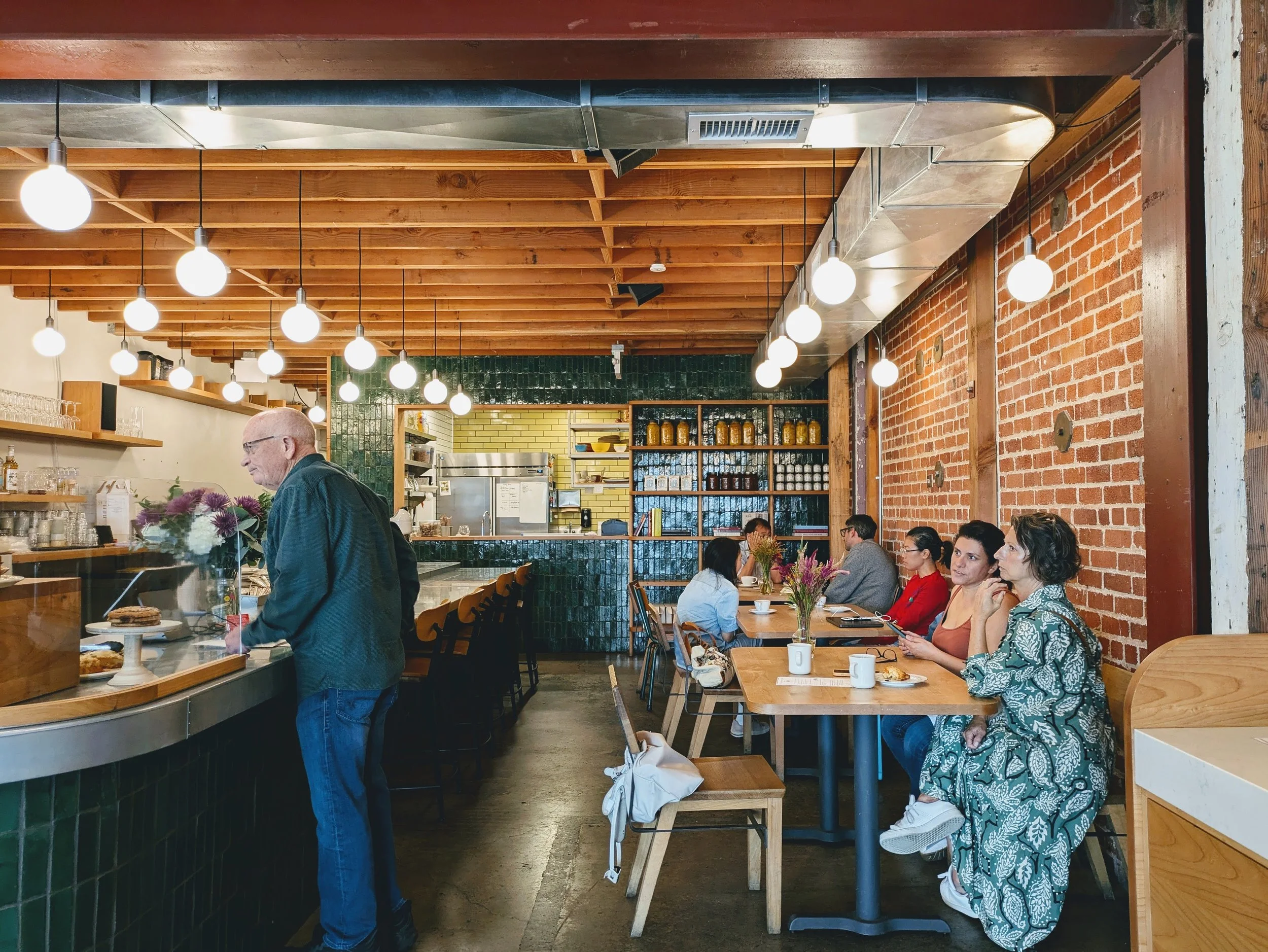Evergreen Cafe_Berkeley, CA_A cozy coffee shop interior with wooden ceiling, green tiled counter, exposed brick walls, and warm lighting. Customers are seated at tables, and a barista is preparing drinks behind the counter.
