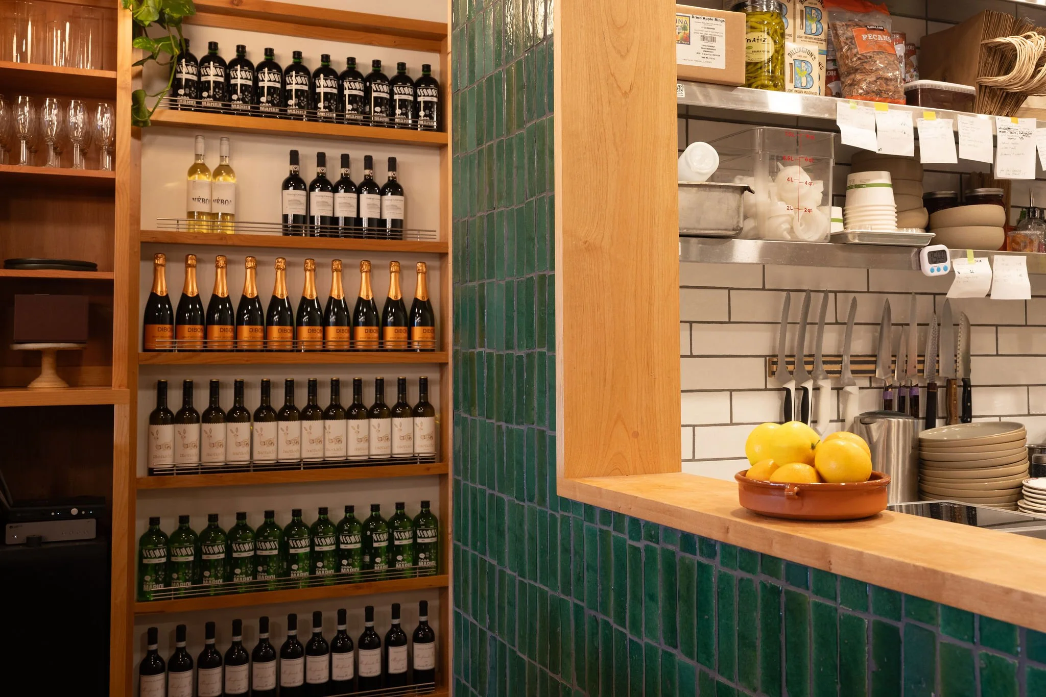 Evergreen Cafe_Berkeley, CA_Part of a restaurant's interior showing a wooden shelf with bottles of wine, bottles of sparkling wine, and bottles of beer, next to a kitchen counter with lemons in a clay bowl and a tile wall with knives and dishes.