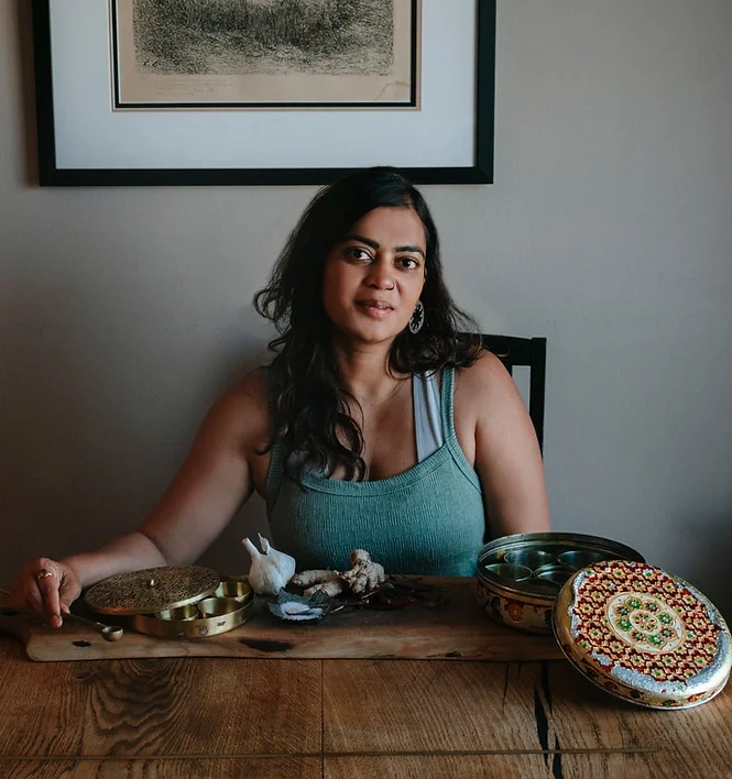A young Indian woman wearing a turquoise tank top sits at a wooden table filled with spice jars, garlic and ginger.
