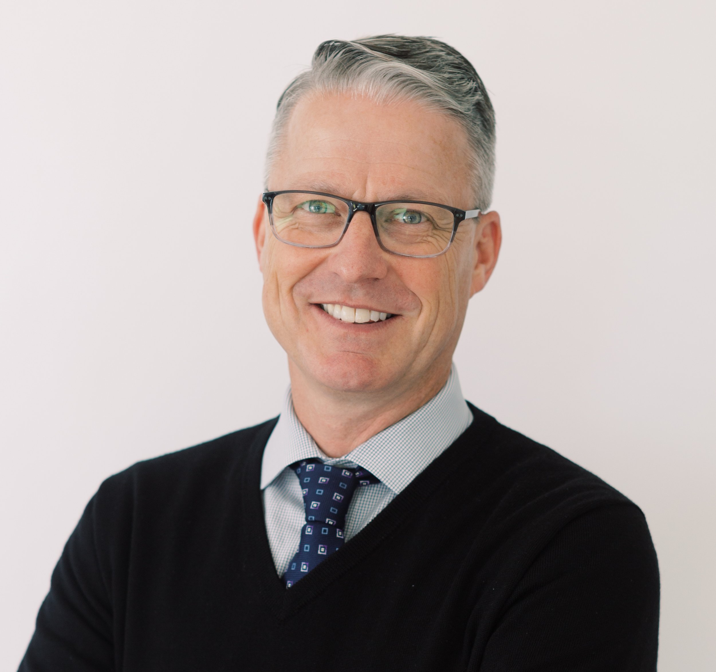 Close-up of a smiling middle-aged man with gray hair, wearing glasses, a dark sweater, a collared shirt, and a patterned tie, against a plain white background.
