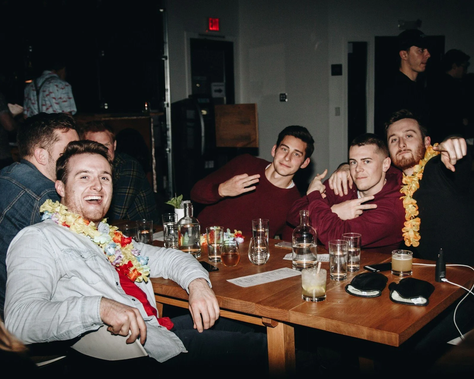 Group of friends sitting at a table in a dimly lit indoor setting, enjoying drinks and wearing leis, with some smiling and making gestures, in what appears to be a lively social gathering or party.