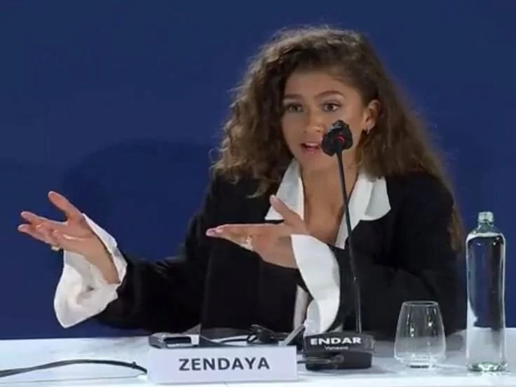 Woman with curly brown hair wearing a black blazer with white cuffs, speaking into a microphone at a panel discussion, seated at a table with a nameplate reading 'ZENDAYA,' a glass of water, and a water bottle in front of her.
