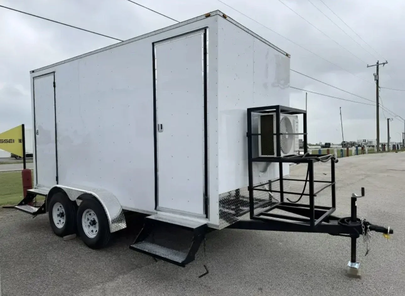A white mobile trailer with dual axles, a small ramp, and an external air conditioning unit mounted on a black frame, parked on a paved lot under cloudy skies.