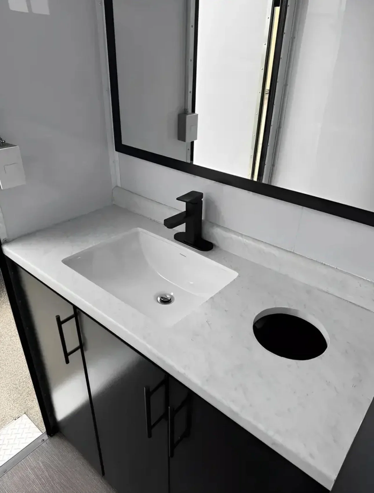 White bathroom sink with black faucet and cabinet below, large mirror above, and a soap dispenser on the wall, with a hole in the sink for a faucet or fixture.