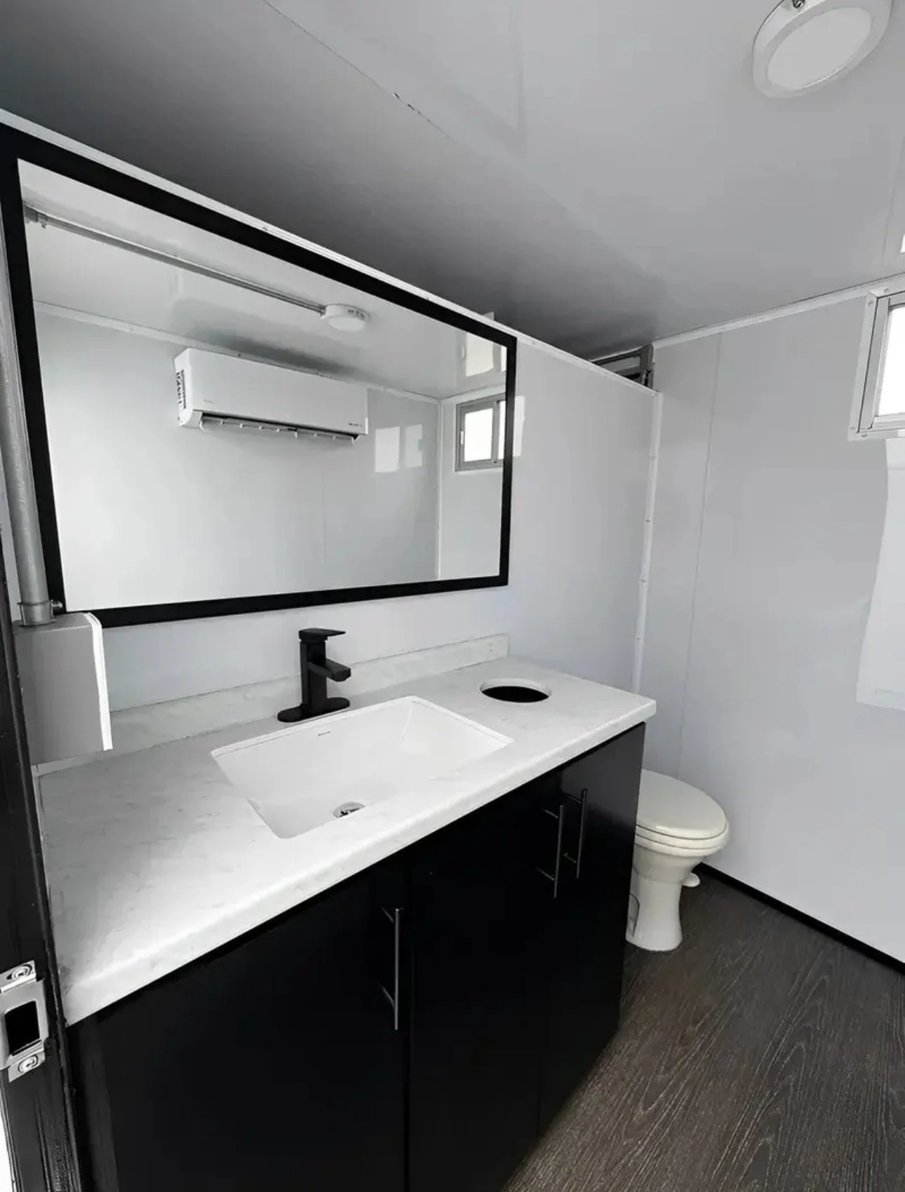 Modern bathroom with a large mirror, black faucet, white sink, black vanity, and a toilet, with a small window and an air conditioning unit on the wall.