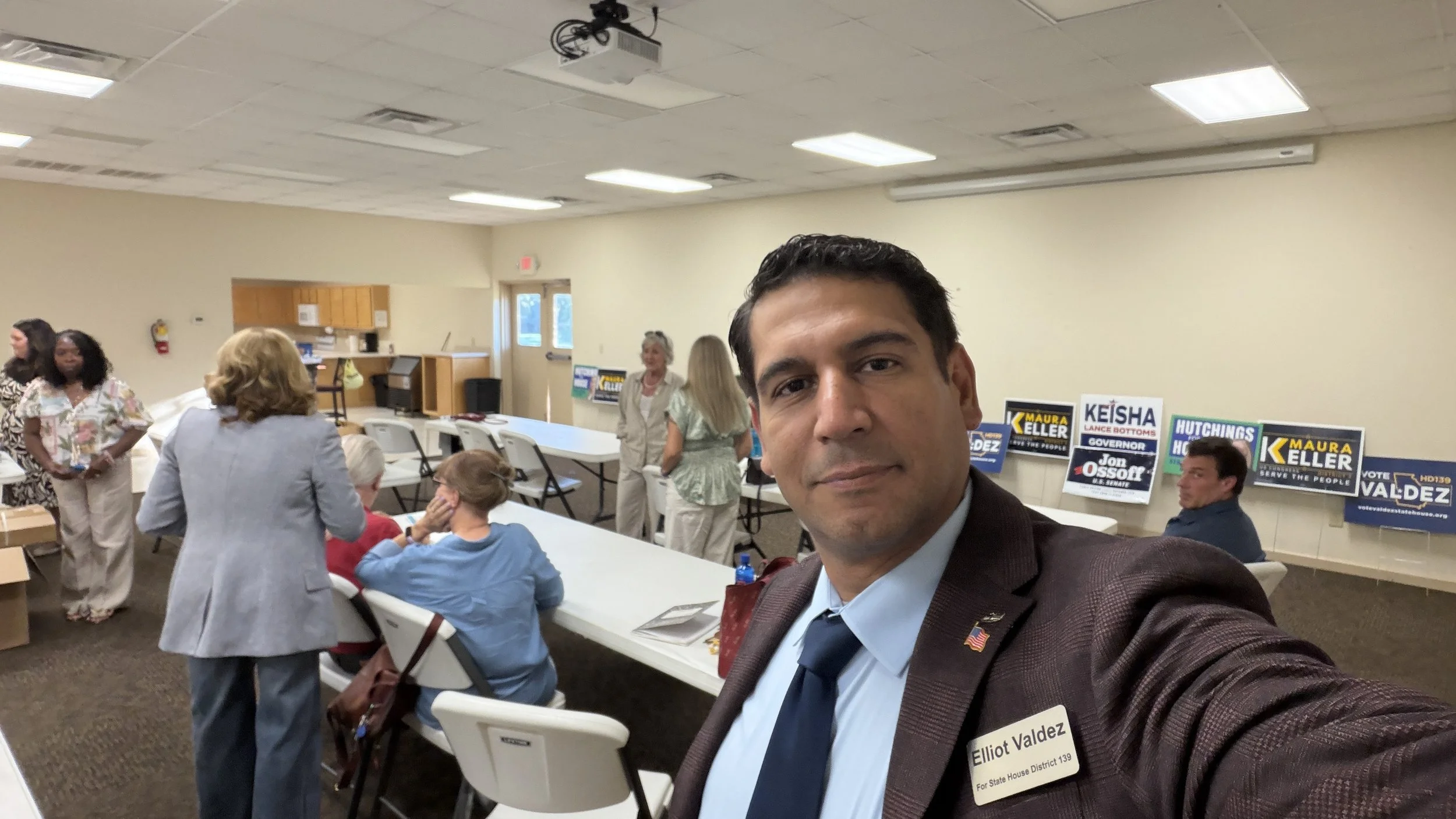 Elliot Valdez taking a selfie at a gathering with several people in the background, including campaign signs for Maura Keller, Keisha Lance Bottoms, and other political candidates.