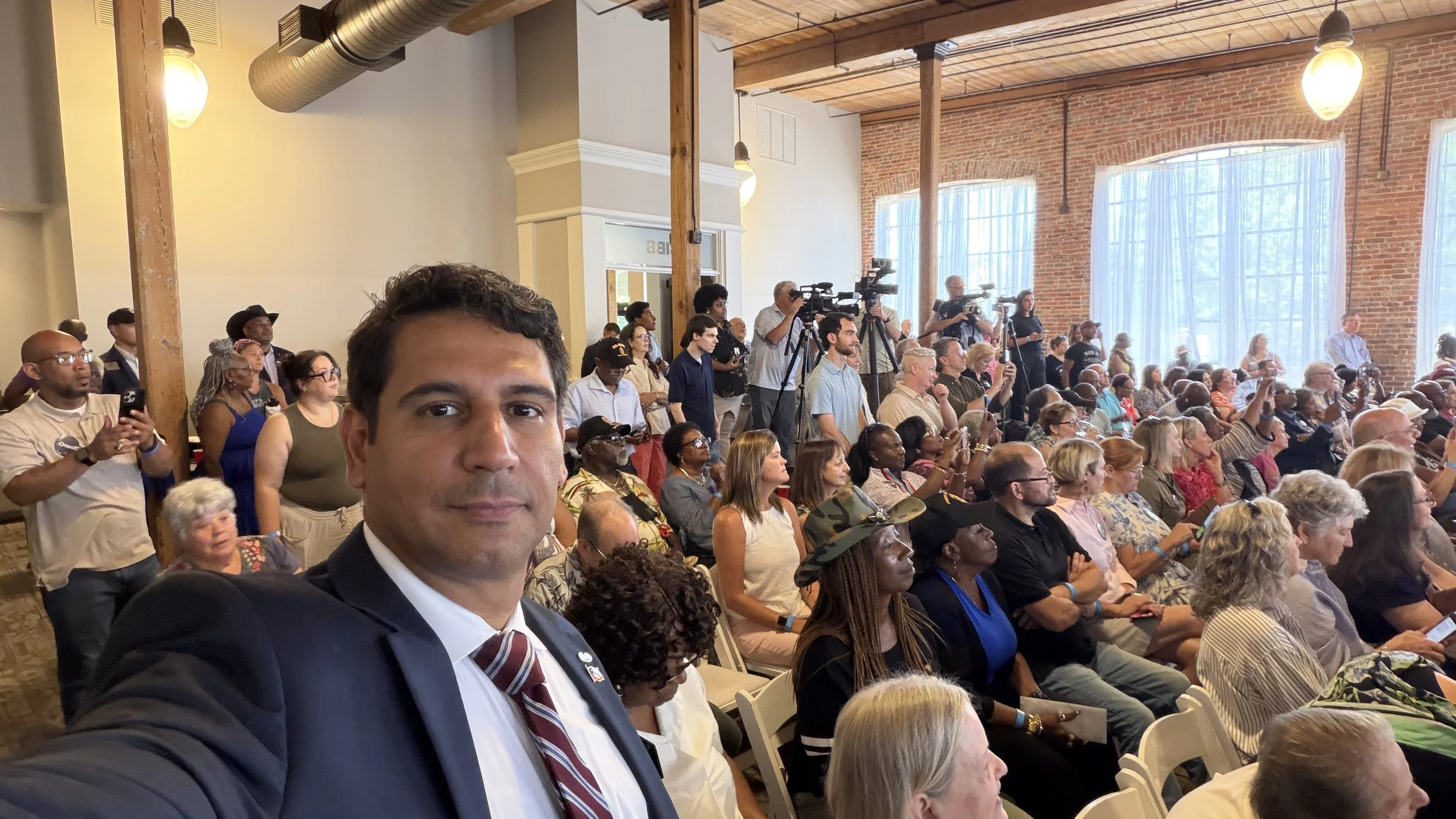 Elliot Valdez in a suit and red-striped tie taking a selfie at a large indoor event with many seated and standing people, some with cameras, in a venue with high ceilings, exposed brick walls, large windows, and wooden beams.