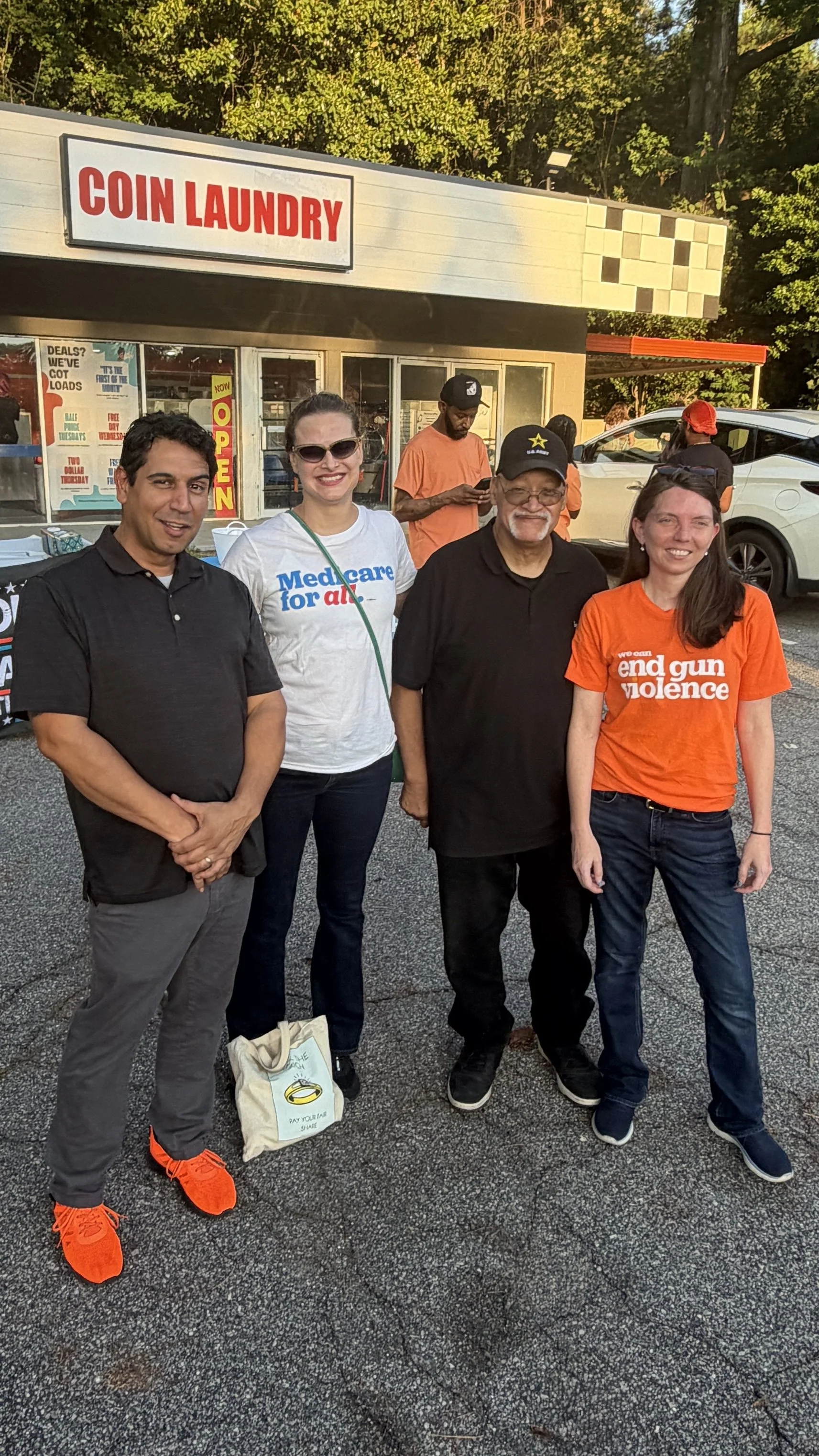 Four people standing outside a coin laundry building during daytime, smiling for the photo. Two women and two men. One woman is wearing sunglasses and a white t-shirt that says 'Medicare for all.' The other woman is in an orange t-shirt that says 'we can end gun violence.' One man has a black cap and glasses, and the other man is wearing bright orange running shoes.