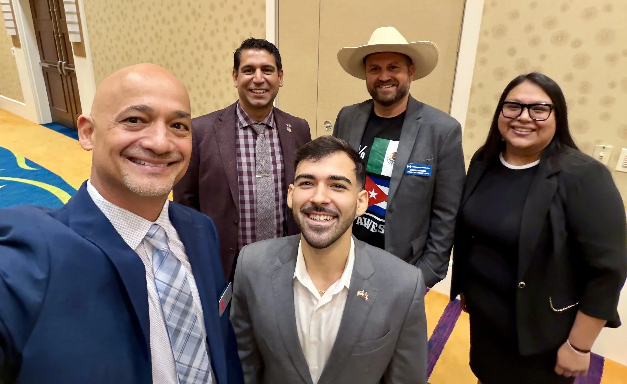 Group of six smiling people taking a selfie inside a conference center with patterned wallpaper and a blue and yellow carpet.