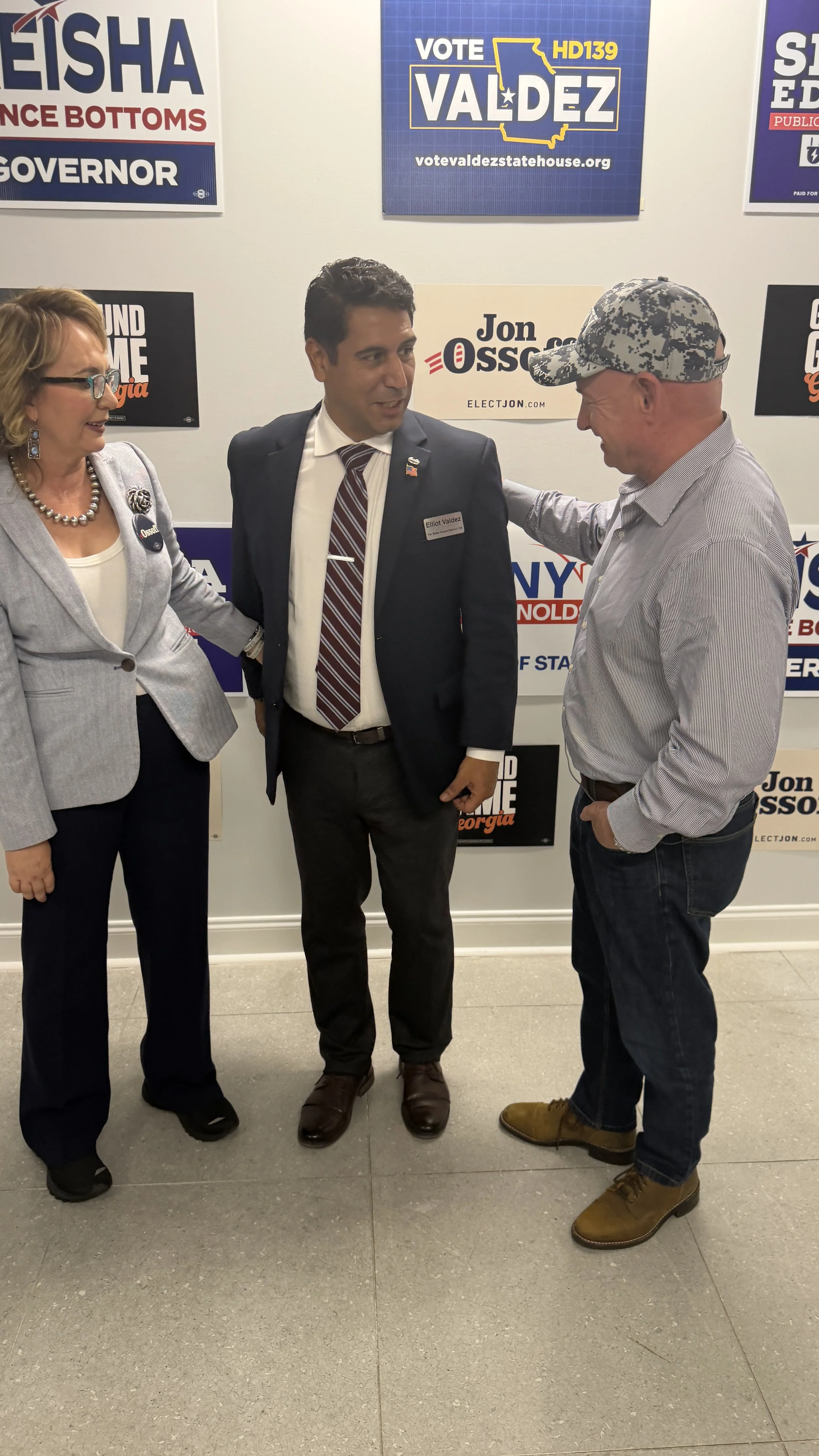 Three people standing together at a political event, in front of campaign signs for Valdez, Jon Ossoff, and other candidates. The woman on the left is wearing glasses, a light blazer, and earrings, and the man on the right is wearing a camouflage cap and a striped shirt. The man in the middle is wearing a suit and tie and is engaged in conversation with the man on the right.