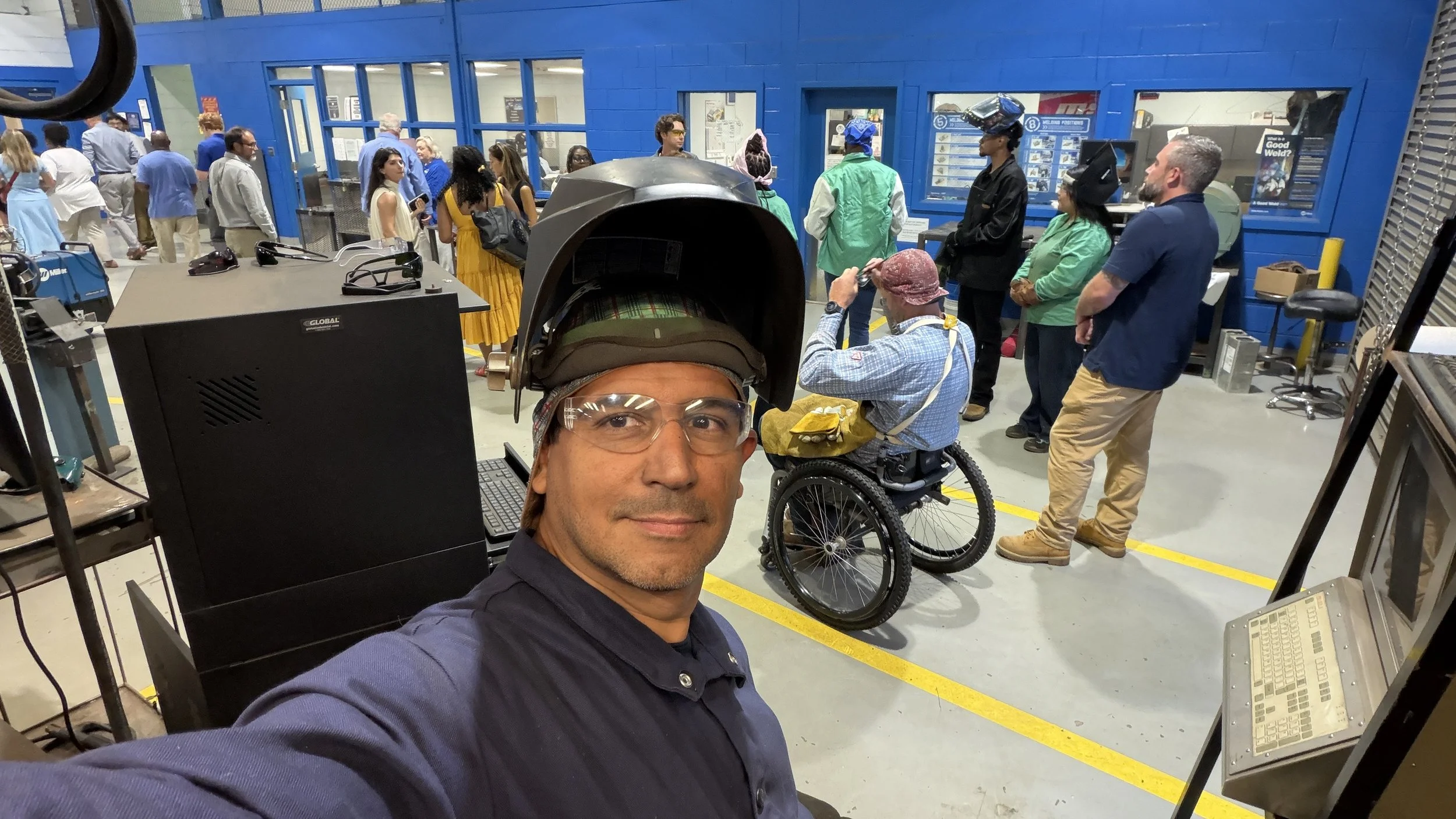 A man wearing glasses and a safety helmet taking a selfie in a building supply store or hardware store, with several people in the background, including a person in a wheelchair and others waiting in line or shopping.