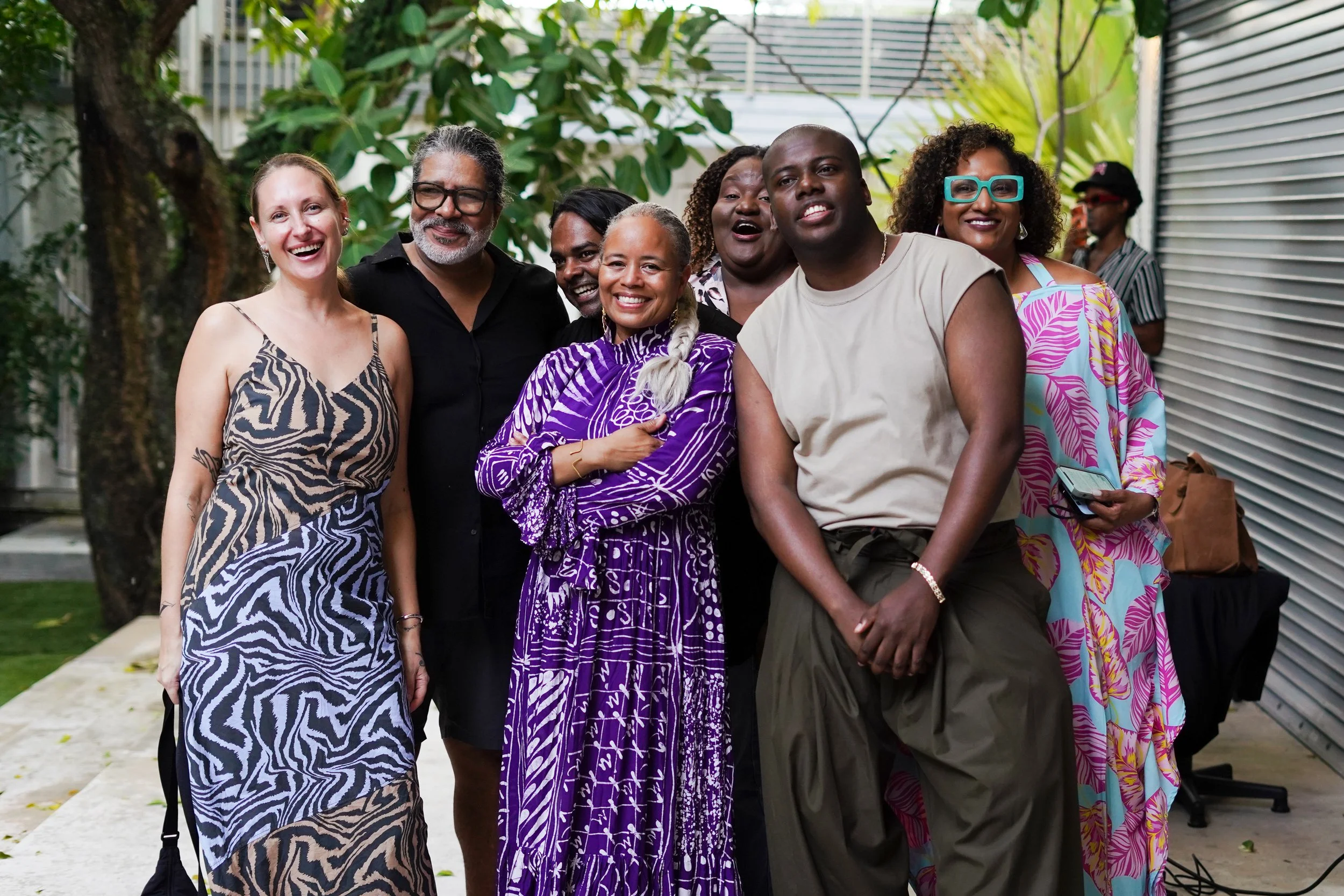 A group of nine diverse people smiling and posing outdoors, with greenery and a metal wall in the background.