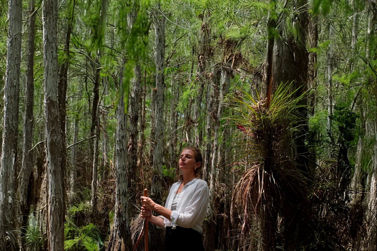 A woman standing in a dense forest with tall trees and green foliage, holding a walking stick, and wearing a white shirt.