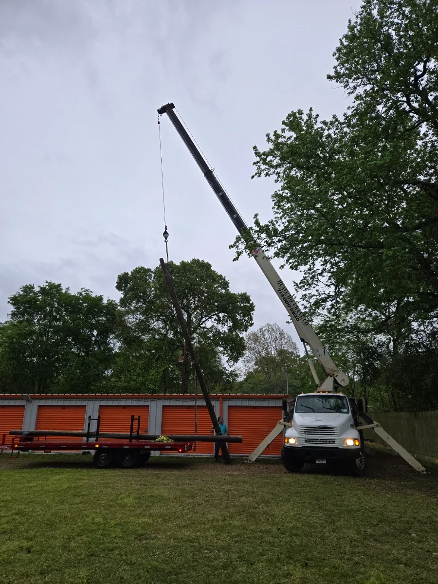 A crane truck lifting a pole or piece of equipment, with a person standing nearby holding a cable or rope, in a yard with green grass, trees, and orange storage units or containers in the background.