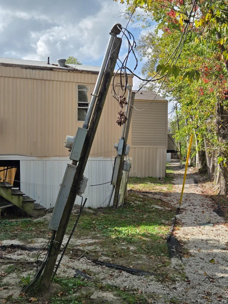 Upside-down utility poles leaning against a house with electrical transformers and wires, next to a pathway and trees.