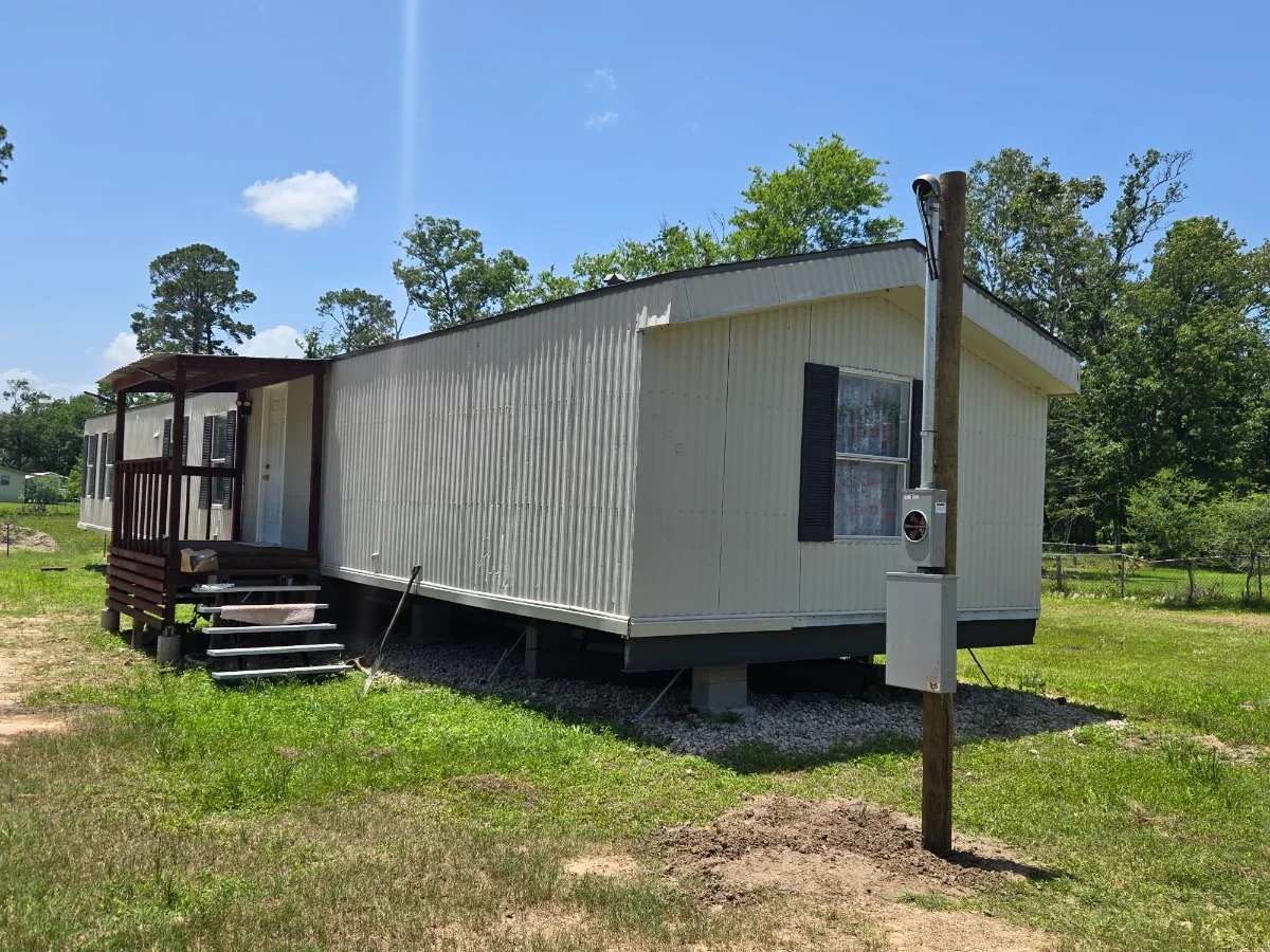 Mobile home with small porch, steps, window with shutters, and utility meter, surrounded by grass and trees under a blue sky.