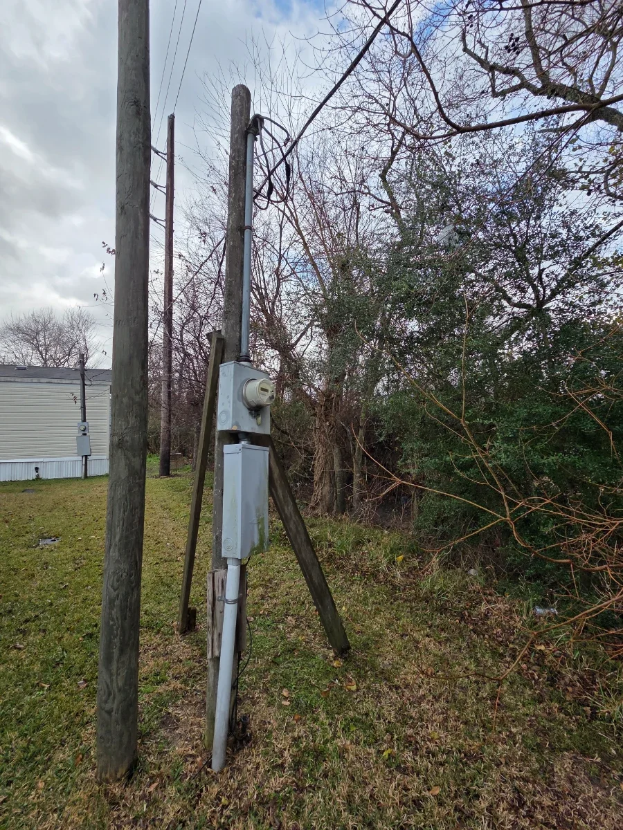 Electric utility pole with a weathered wooden support leaning against it, electrical wires running overhead, and some trees and a building in the background under a cloudy sky.