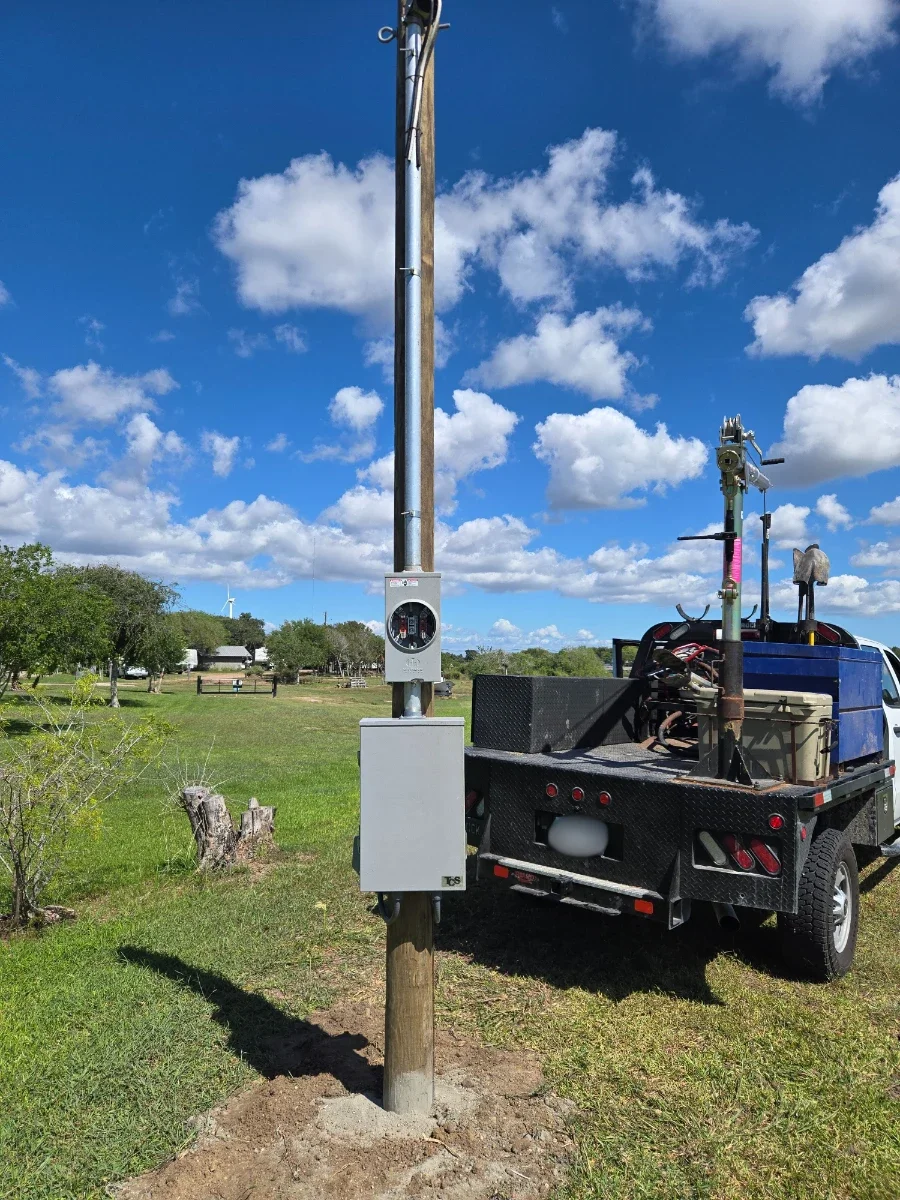 A utility truck parked next to a wooden utility pole with electrical equipment and meters installed, set in a grassy field with trees and a partly cloudy sky in the background.