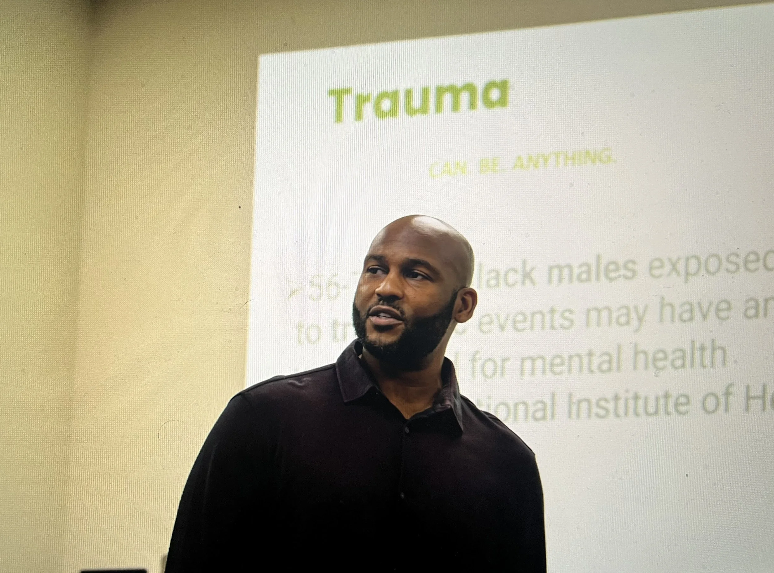 A man with a beard and bald head standing in front of a projected presentation slide titled 'Trauma' with the subtitle 'Can be anything'. The slide contains additional text about the exposure of black males to traumatic events and mental health.