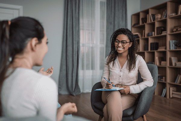 A woman with long brown hair sitting on a sofa, smiling, during a therapy or counseling session with a therapist who is taking notes on a clipboard.