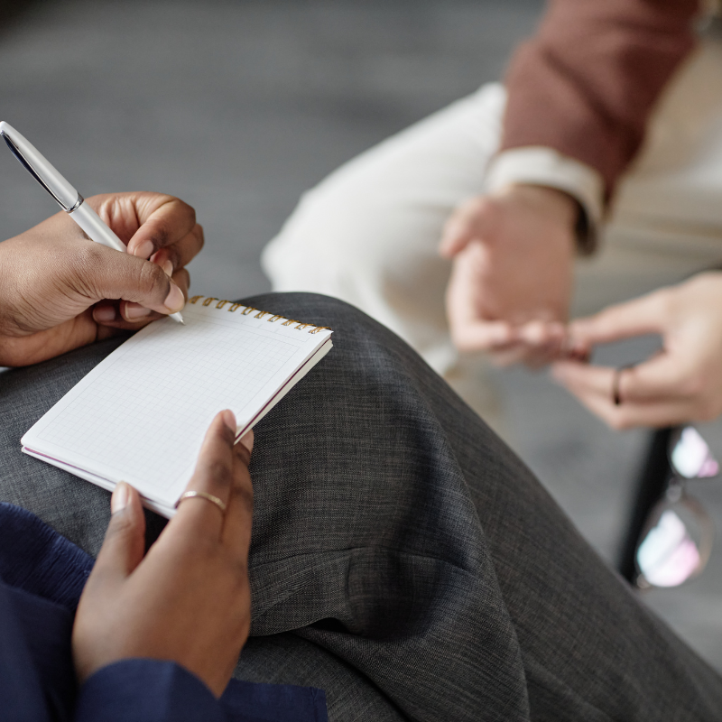A person taking notes in a small notebook with a pen, while another person in the background holds a smartphone, both seated.